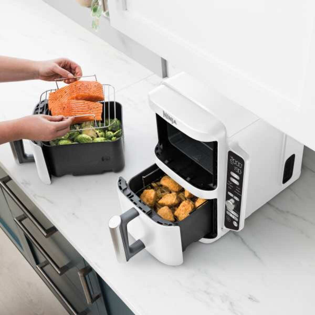 Biała frytkownica powietrzna z jedzeniem w środku. Osoba dodaje jedzenie do frytkownicy.