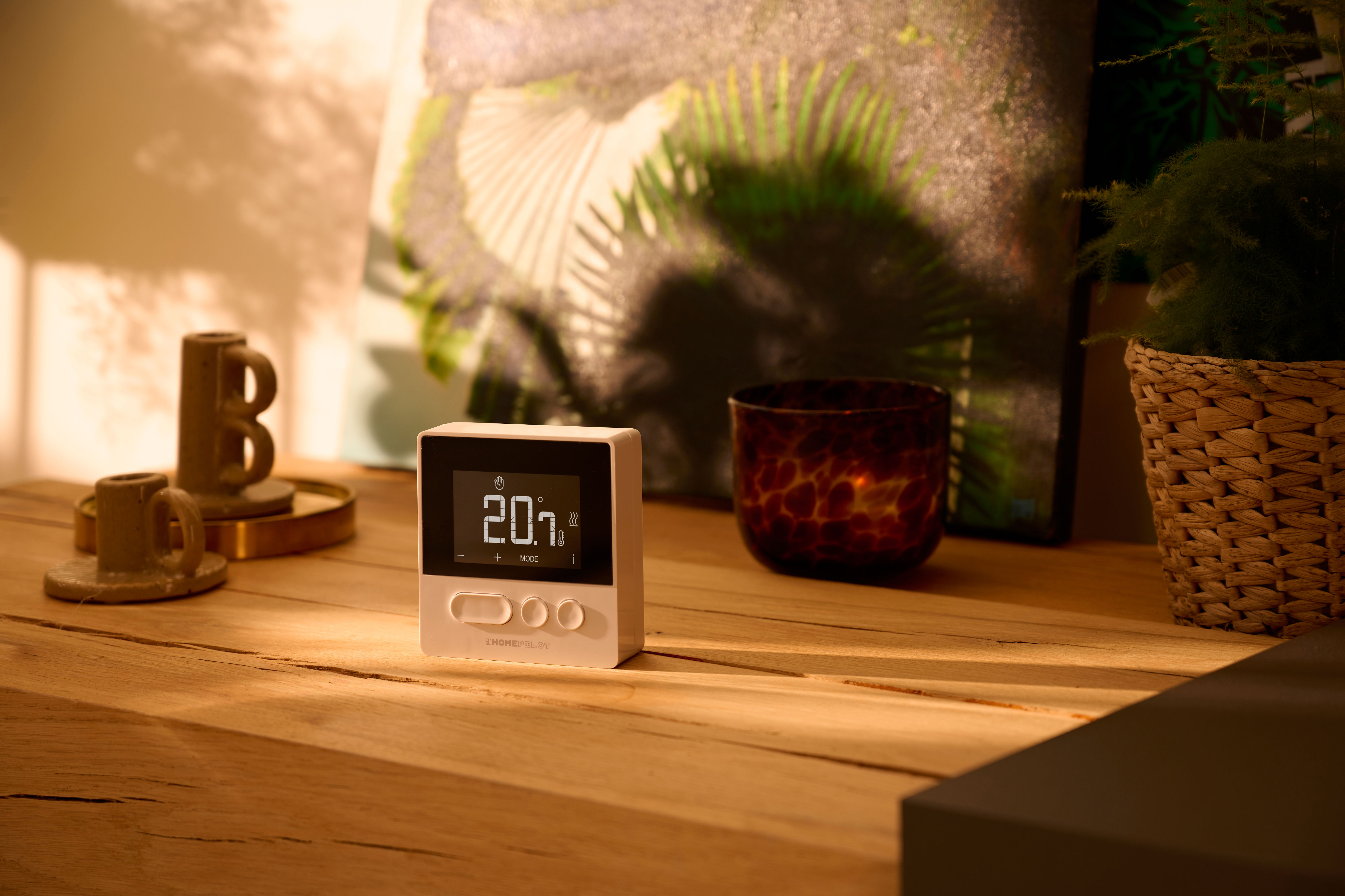 Ein weißer Thermostat auf einer Holzoberfläche mit Dekoration. Ein verschwommener Hintergrund zeigt einen Raum mit Licht.
