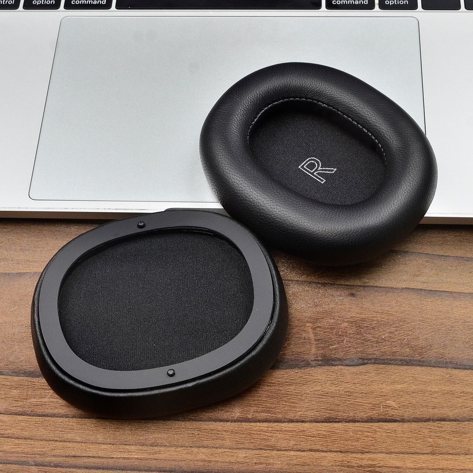 Dwie czarne poduszki nauszne ze znakiem 'R' na jednej, umieszczone na klawiaturze laptopa i drewnianym stole.