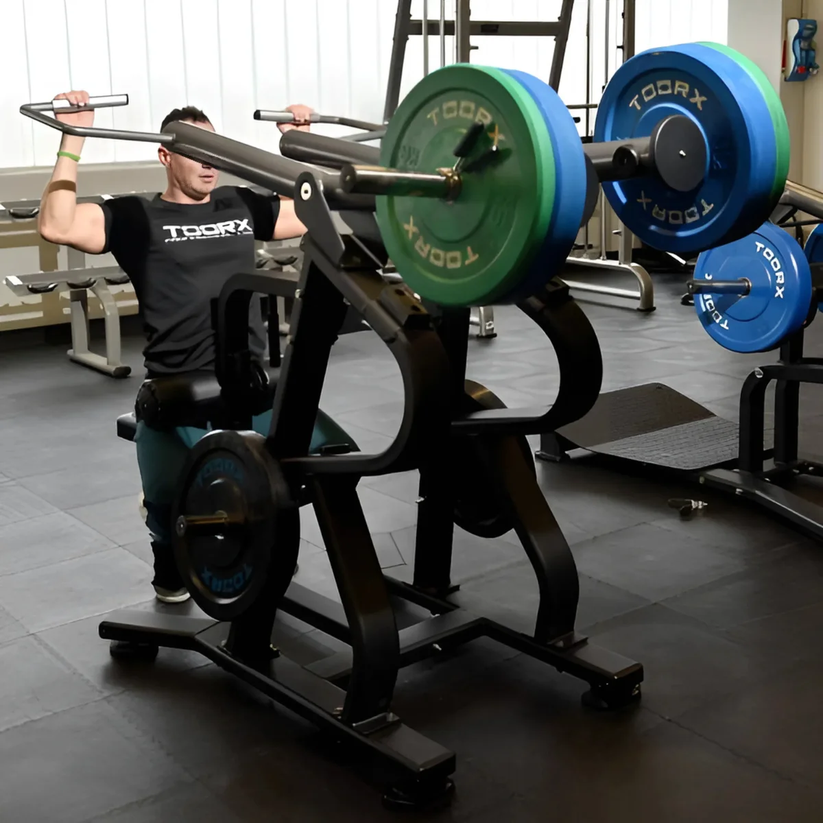 Man in sportschool die een gewichtmachine gebruikt. Blauwe en groene gewichten zijn zichtbaar.