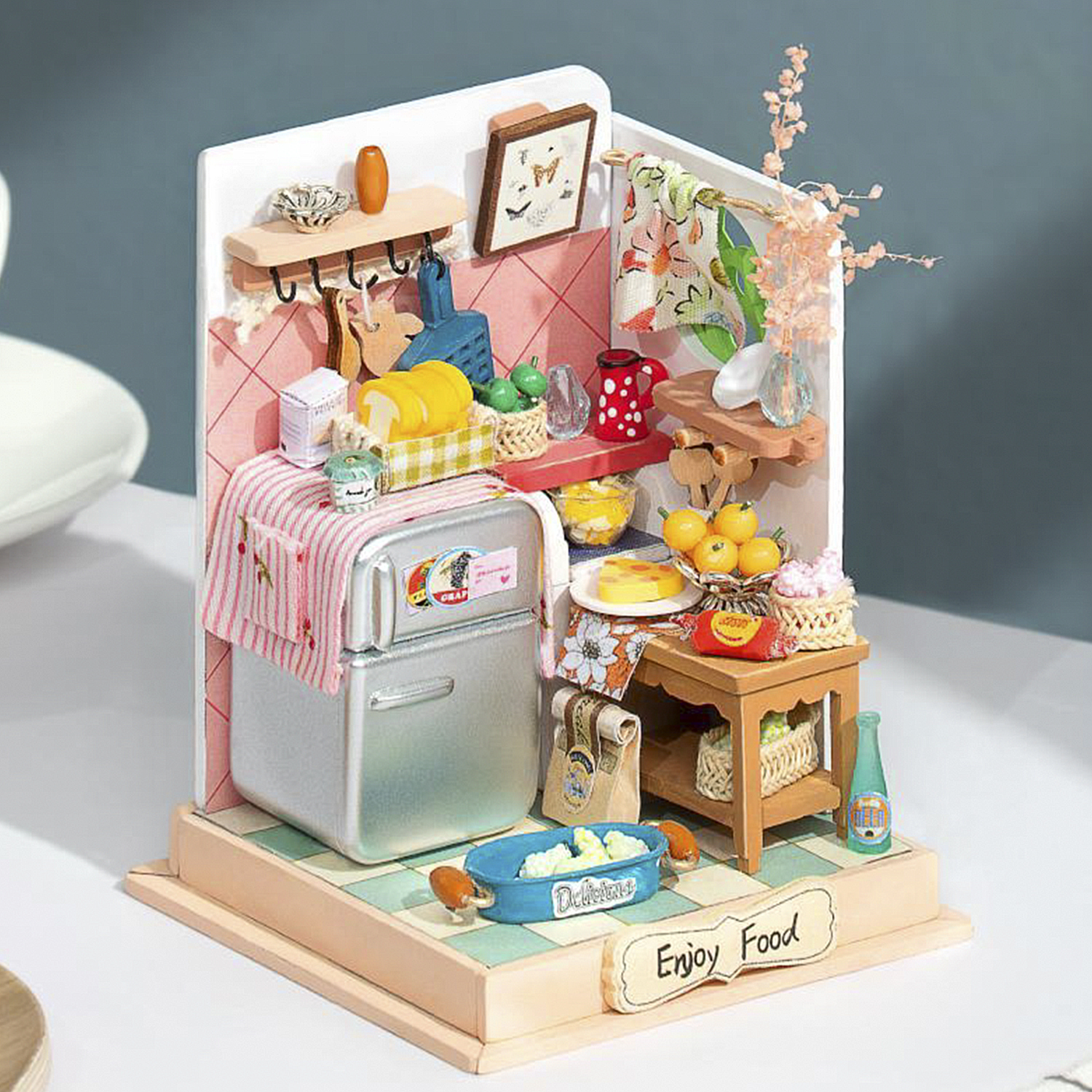 Scena cucina in miniatura con frigorifero, tavolo, cibo e decorazioni. Cartello 'Enjoy Food'.