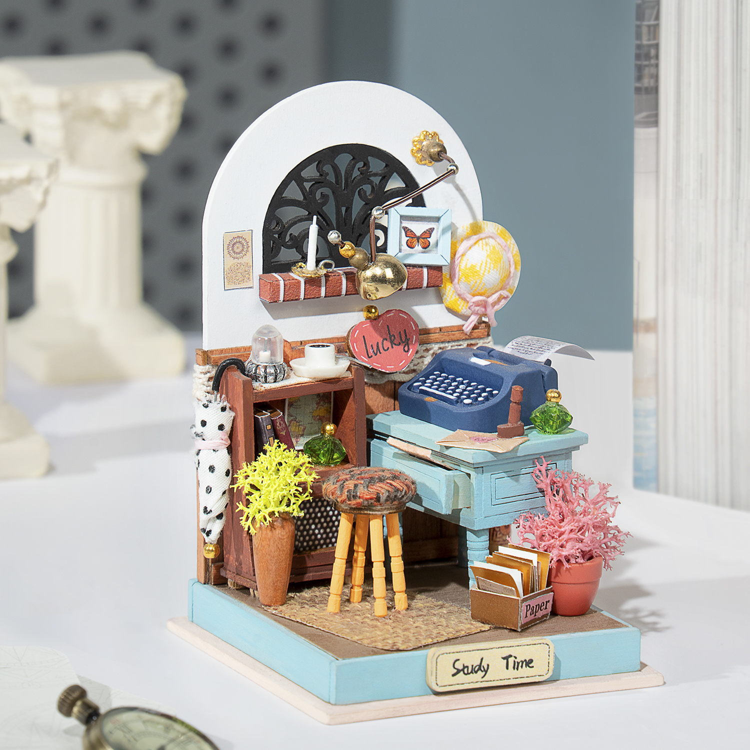 Scena di studio in miniatura, con scrivania, macchina da scrivere, piante e decorazioni. Colori blu e bianco.
