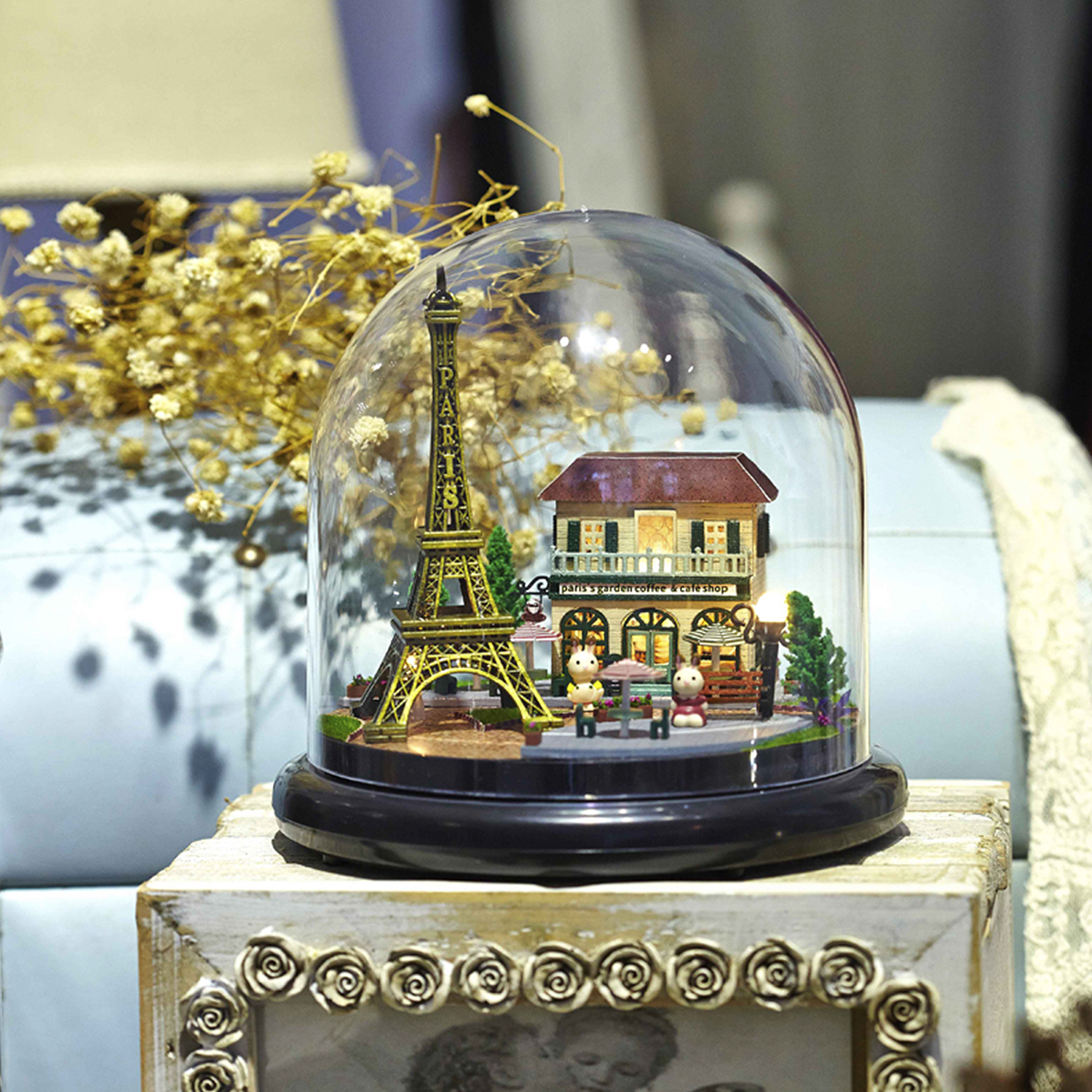 Una miniatura della Torre Eiffel e un modello di negozio all'interno di una cupola di vetro, accanto a un mazzo di fiori secchi su una superficie di legno.