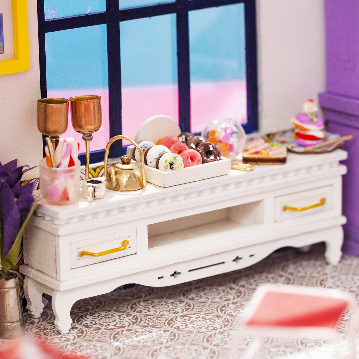 Mobile bianco in miniatura con ciambelle, tazza da tè e dolci davanti a una finestra.