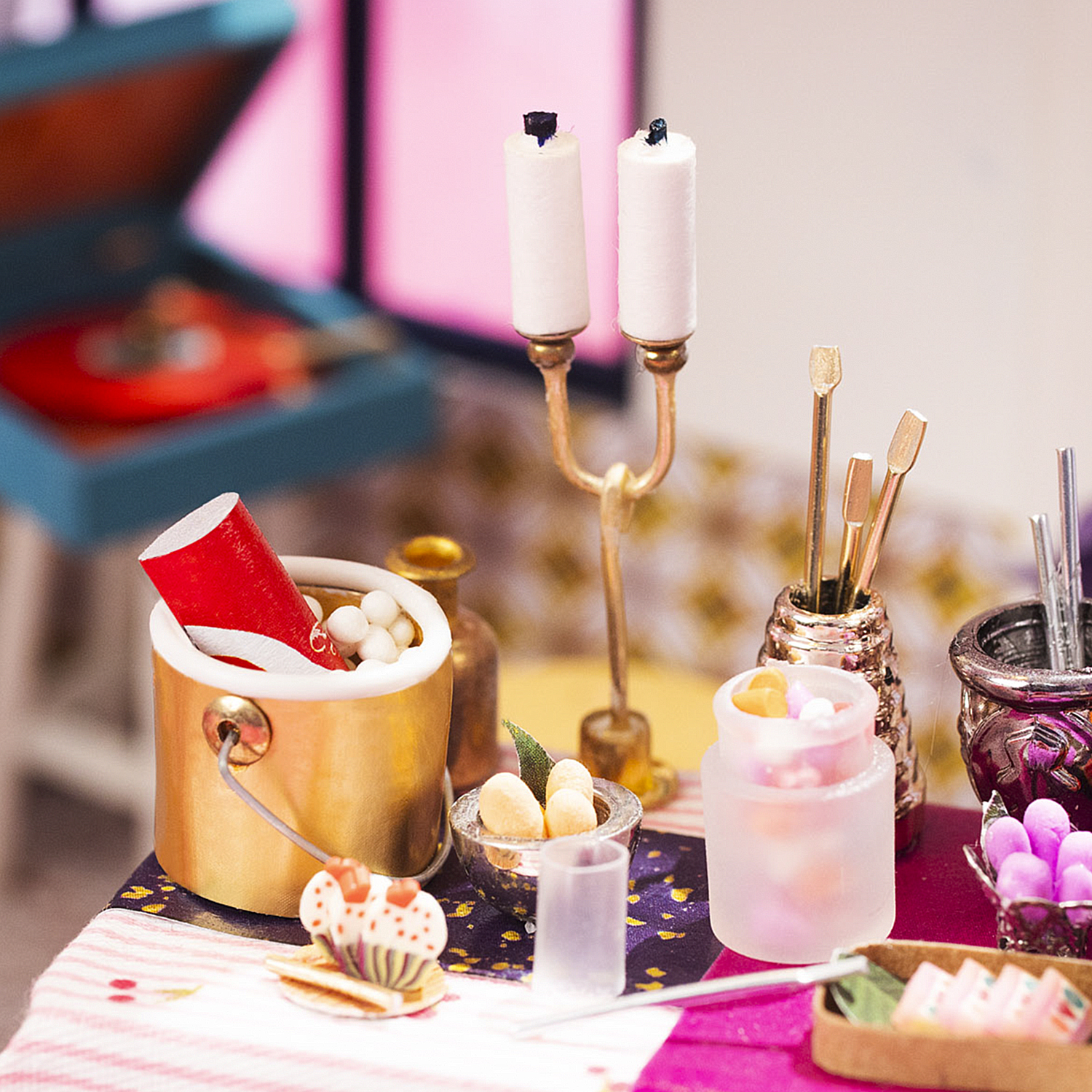 Scena in miniatura di casa delle bambole con candele, dolci e un piccolo giradischi. Accenti rosa e oro.