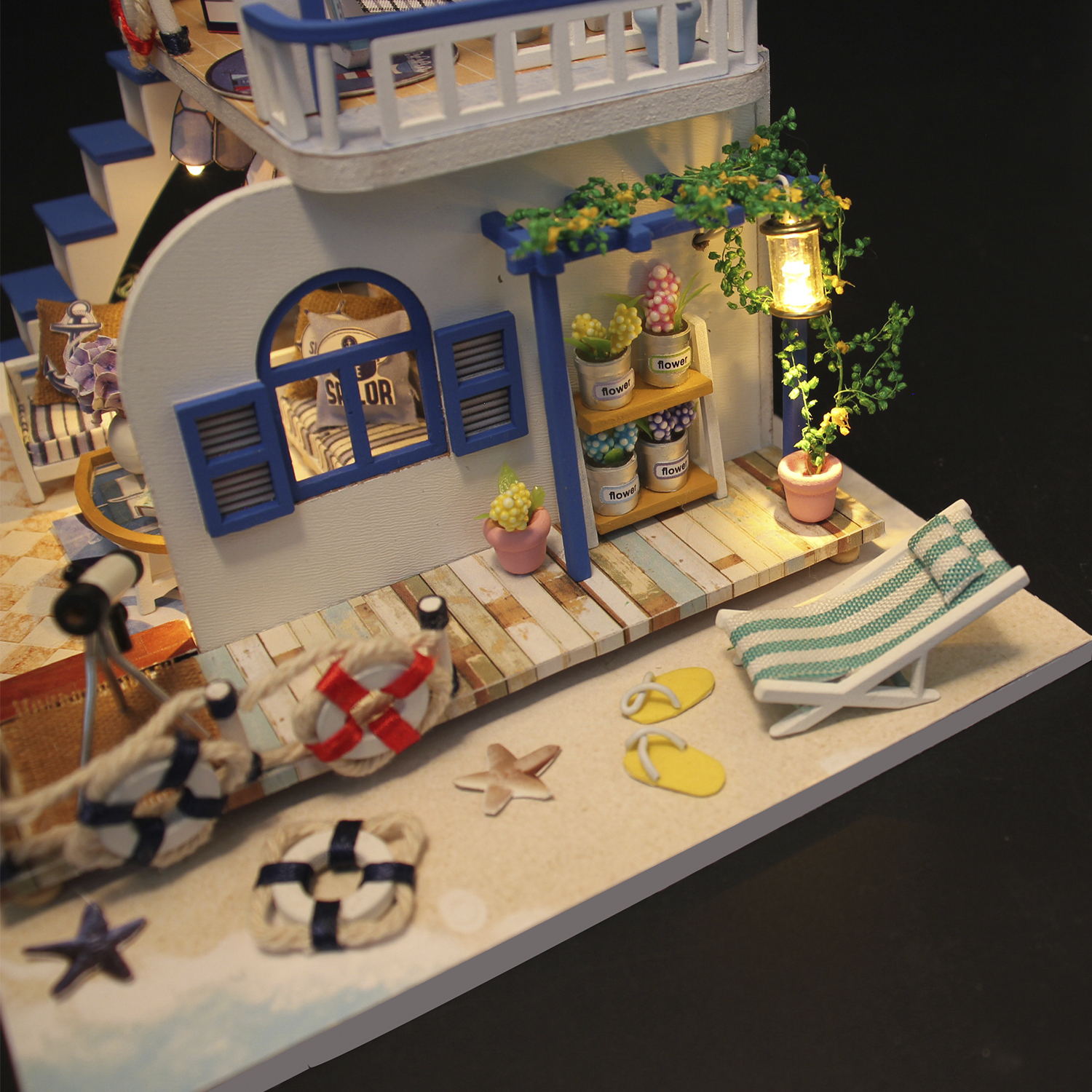 Scena di spiaggia in miniatura con una casa blu e bianca, sedie a sdraio, salvagenti e una piccola palma.