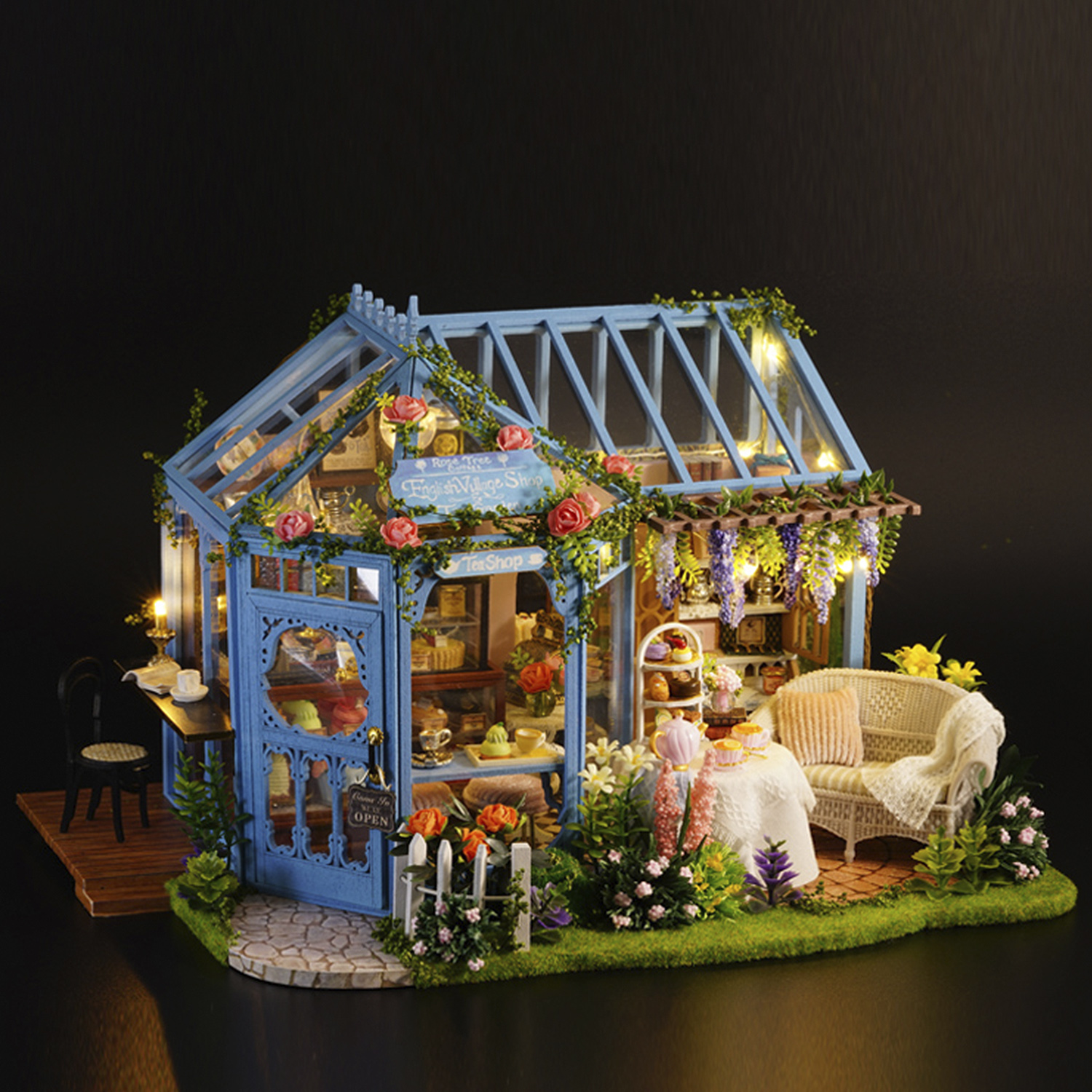 Negozio blu in miniatura con luci fatate e fiori. Dispone di un tavolo con cibo e un divano bianco. Sfondo scuro.