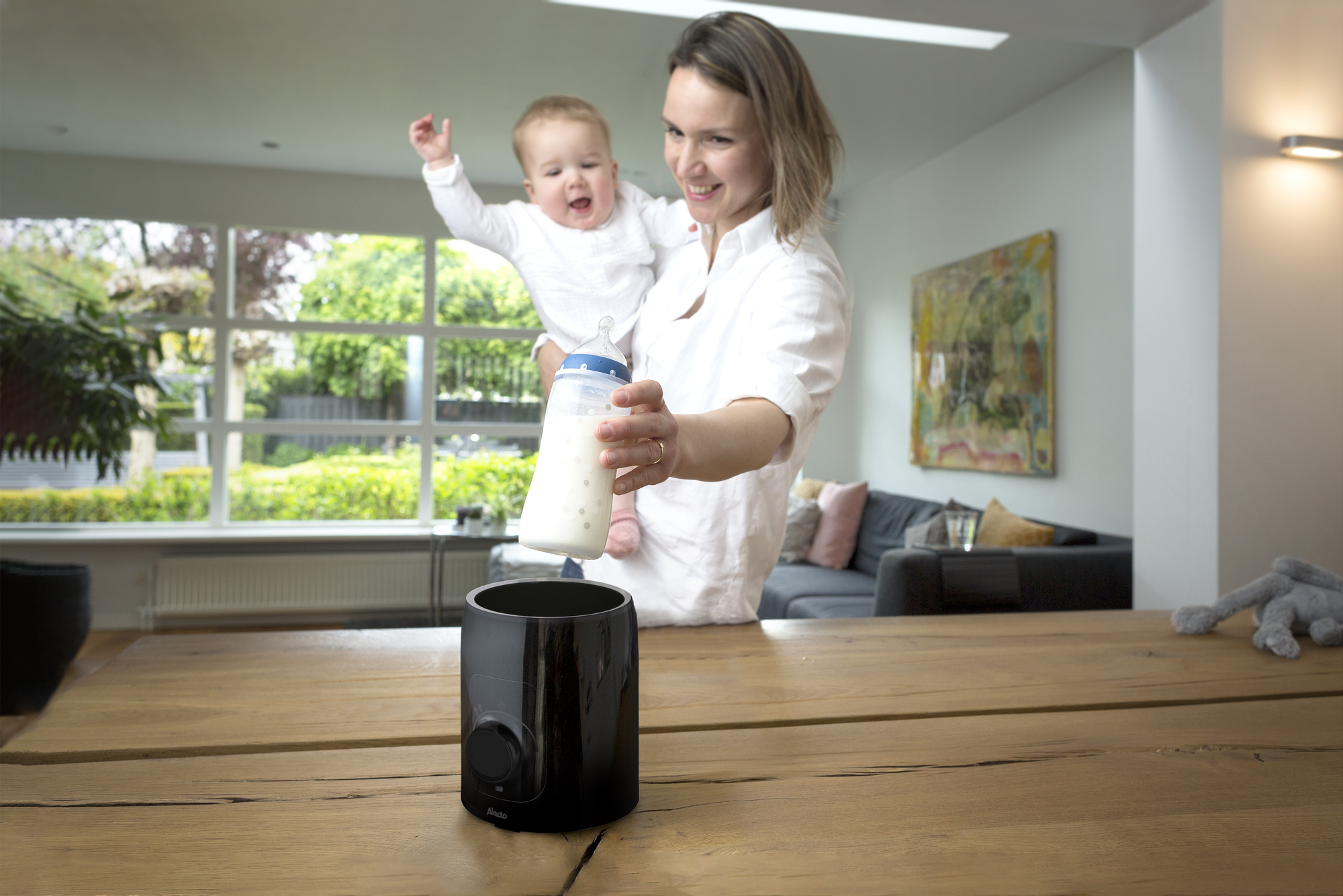 Eine Frau hält ein Baby und eine Flasche Milch. Ein schwarzer Flaschenwärmer steht auf einem Holztisch.
