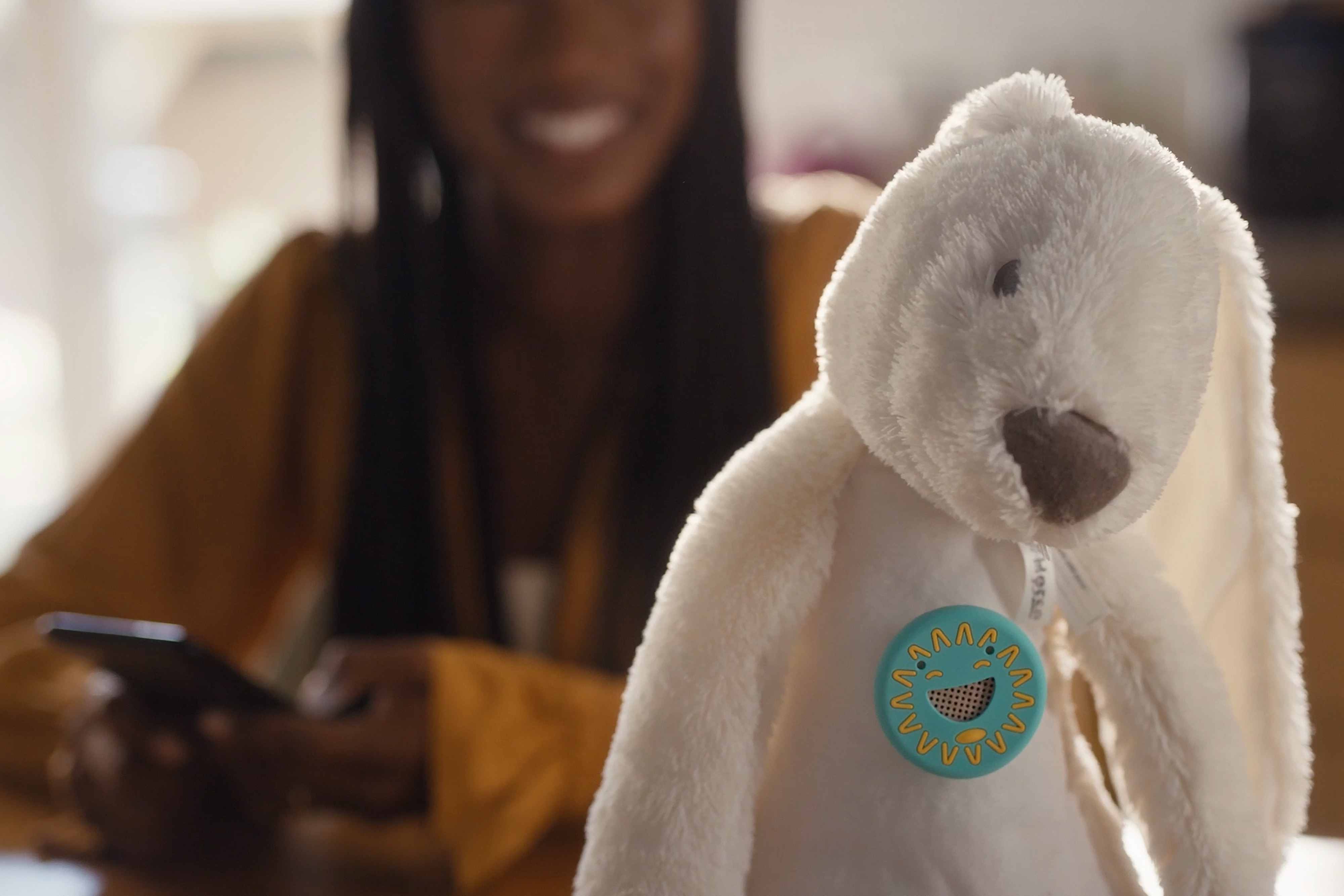 White plush bunny toy with a smiling face, held by a woman using a phone in the blurred background.