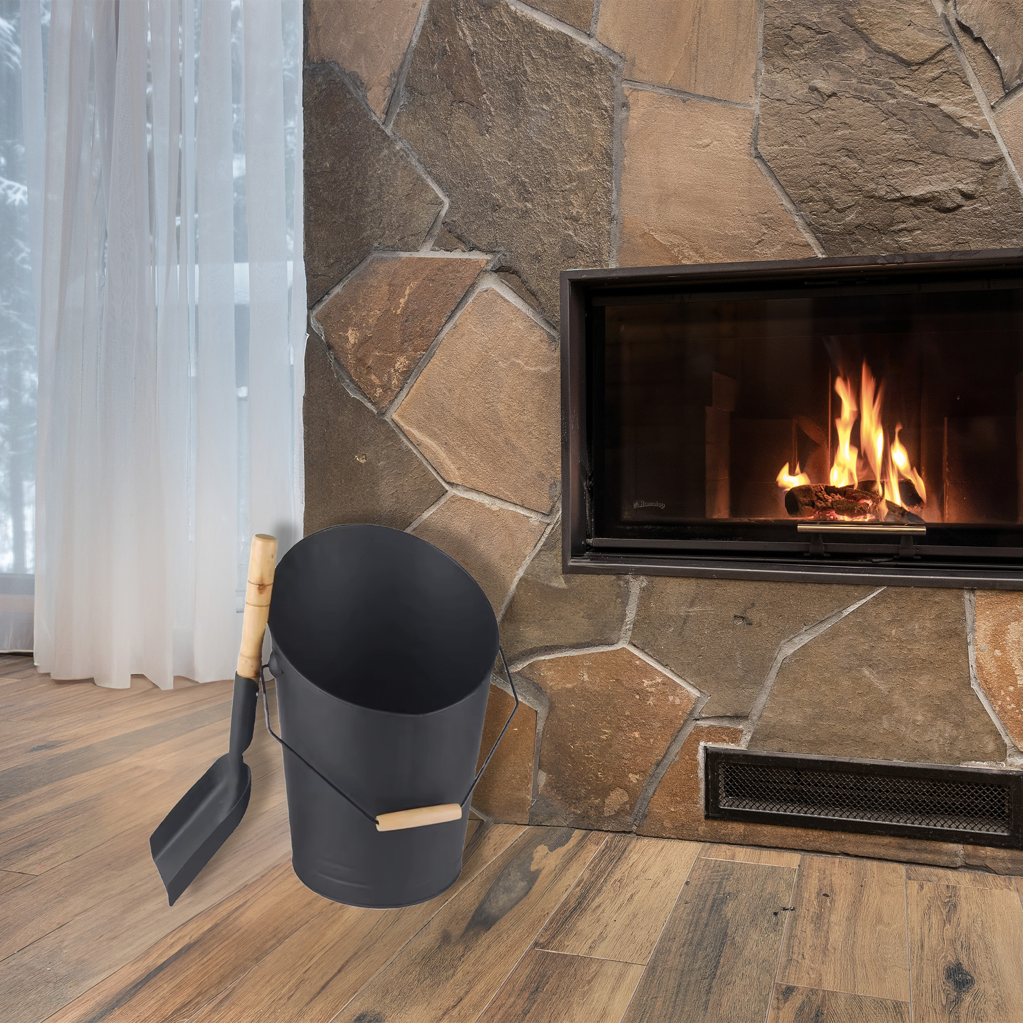 Un cesto de carbón y una pala negros descansan sobre un suelo de madera junto a una chimenea de piedra con fuego.