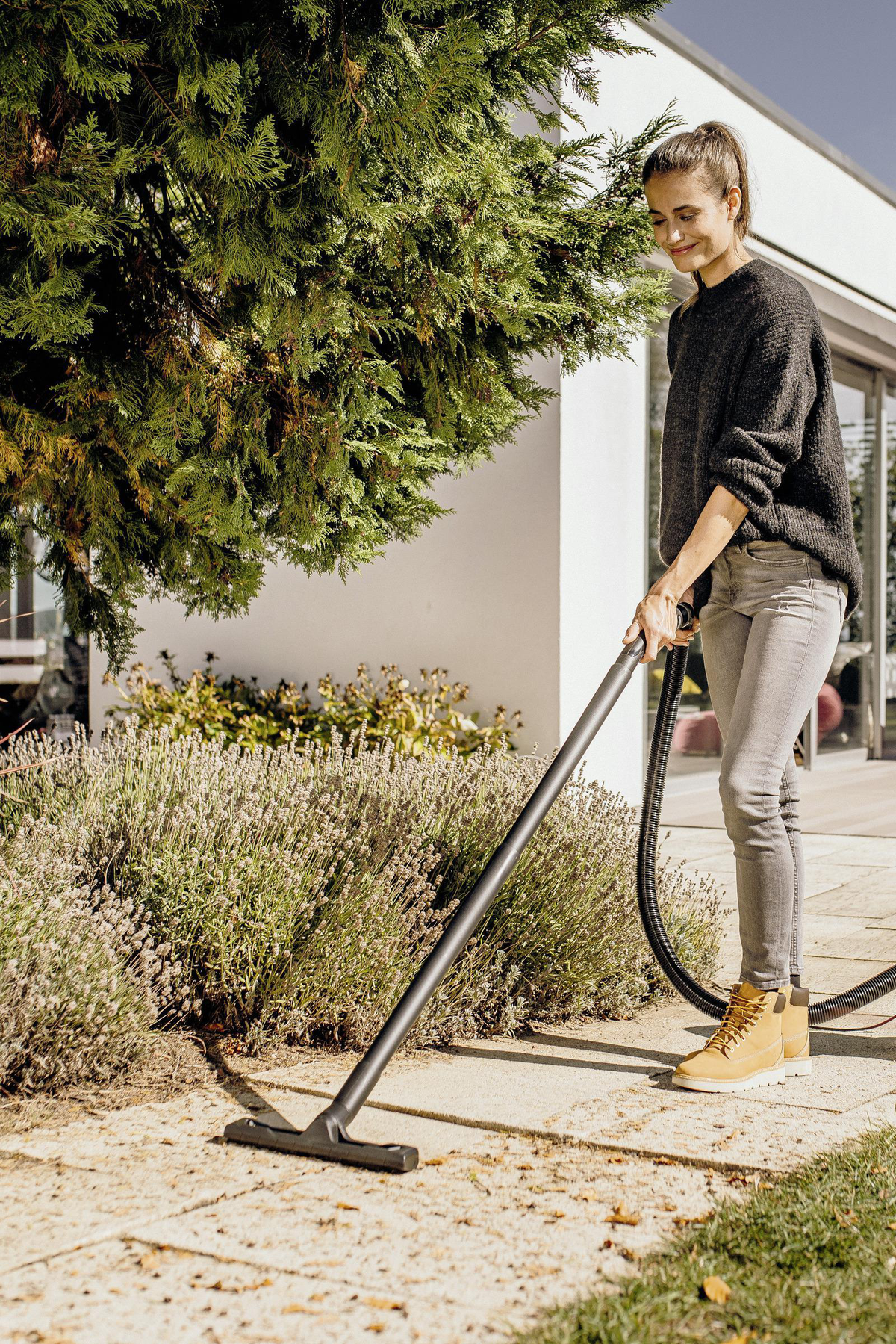 Kobieta używa odkurzacza na patio. Zielone krzewy i dom w tle.