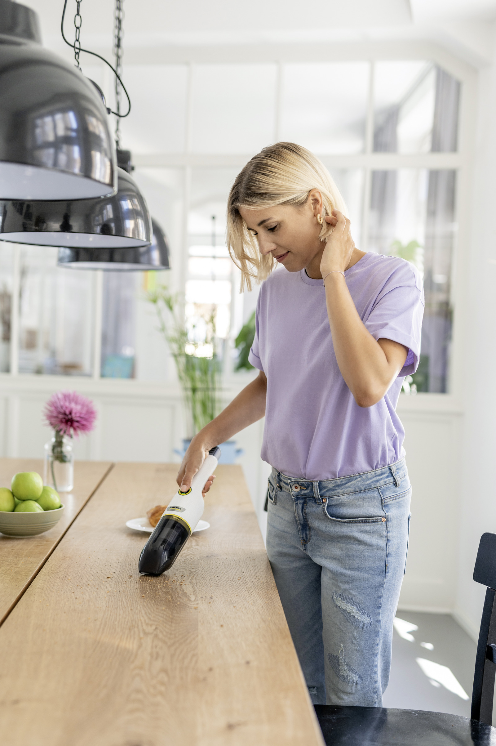 Woman vacuuming crumbs from a wooden table. Pink flower vase, green apples in view.