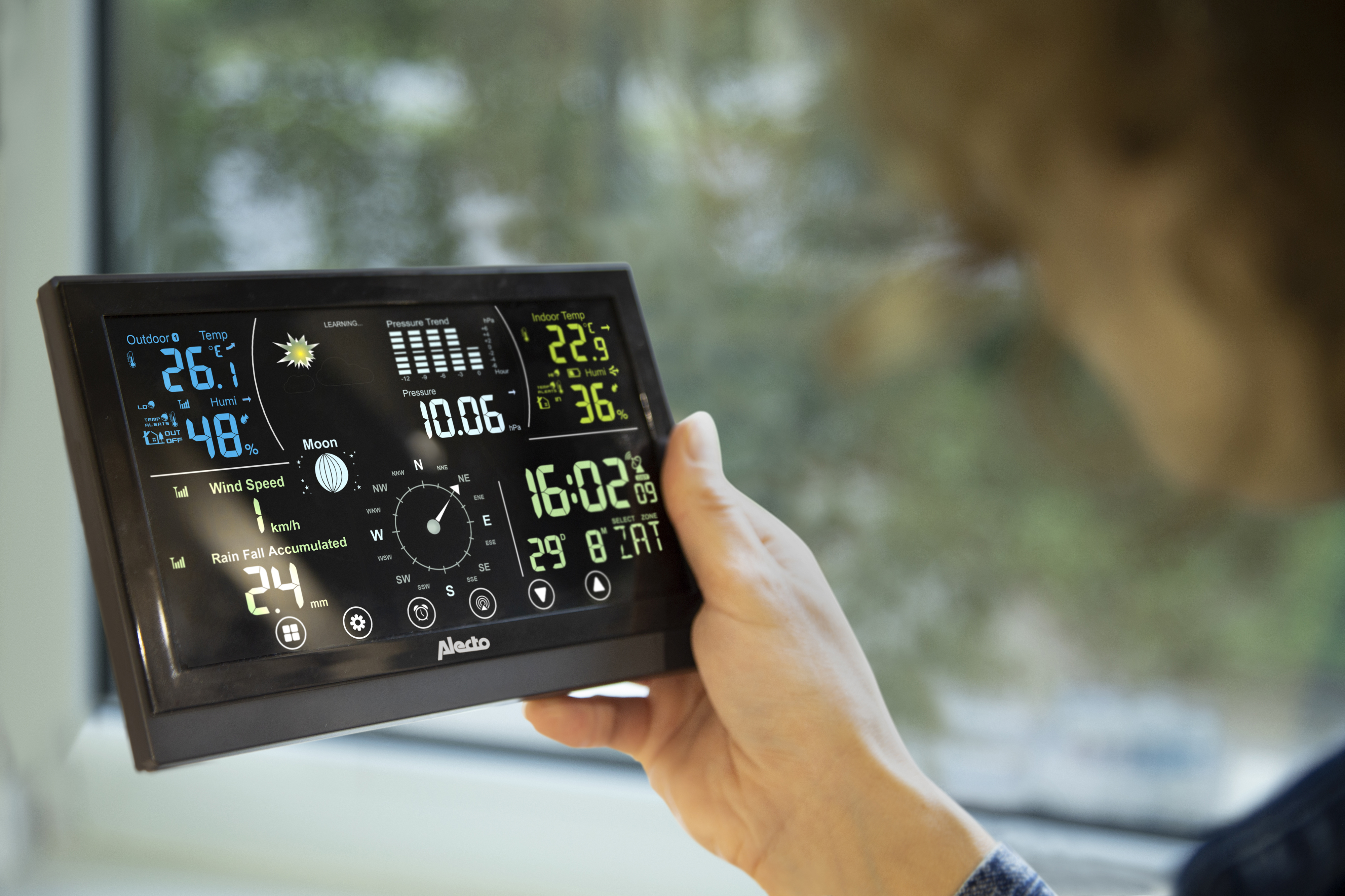 Una persona tiene una stazione meteorologica che mostra temperature esterne/interne, umidità, pressione, velocità del vento e ora.