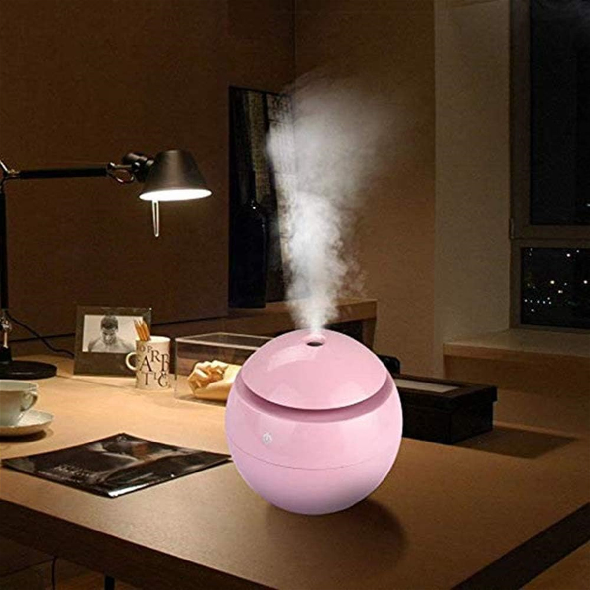 Rosa, kugelförmiger Luftbefeuchter mit weißem Dampf auf einem Holztisch. Lampe und Objekte sichtbar.