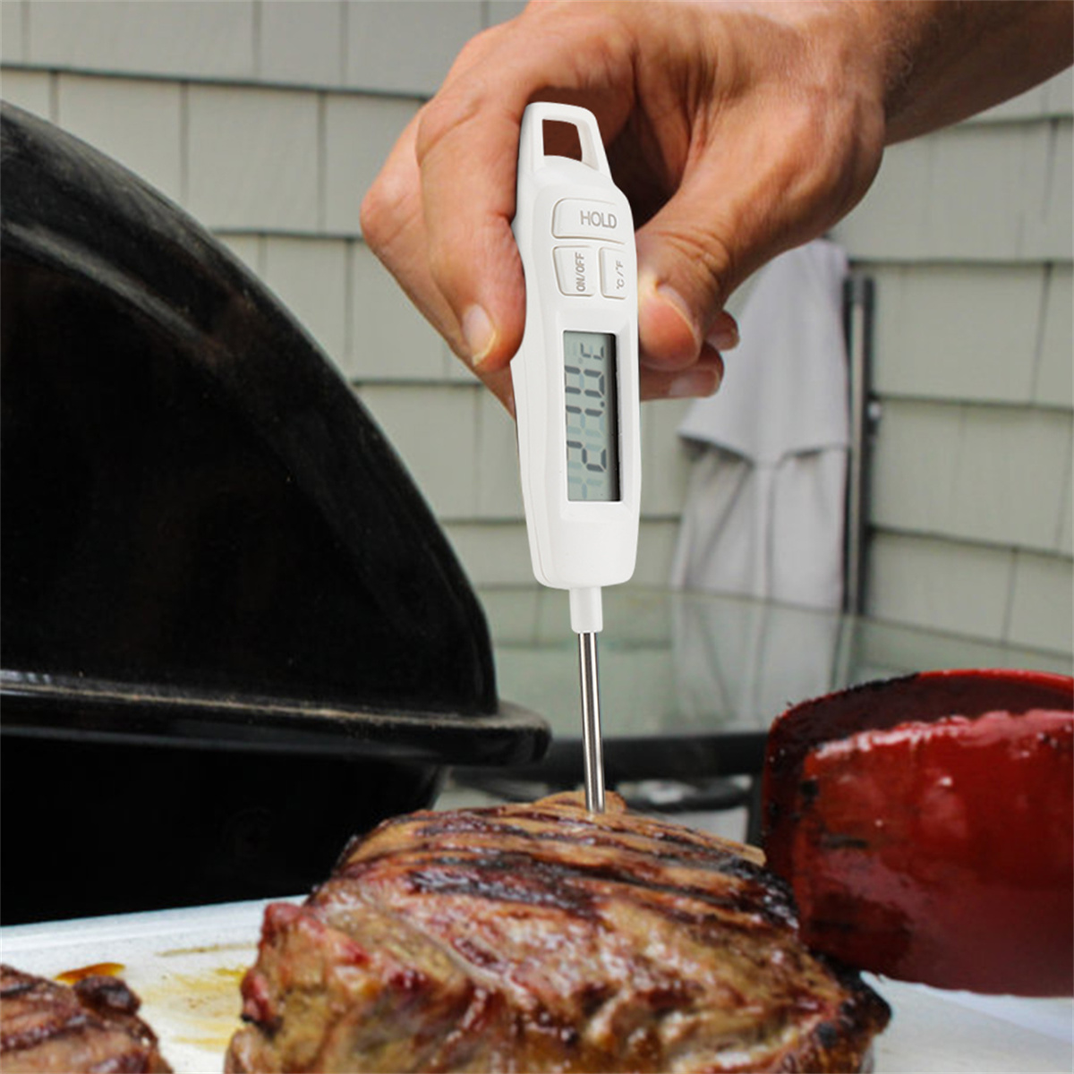 Hand hält weißes Thermometer in Fleisch. Schwarzer Grill und rote Paprika im Hintergrund.
