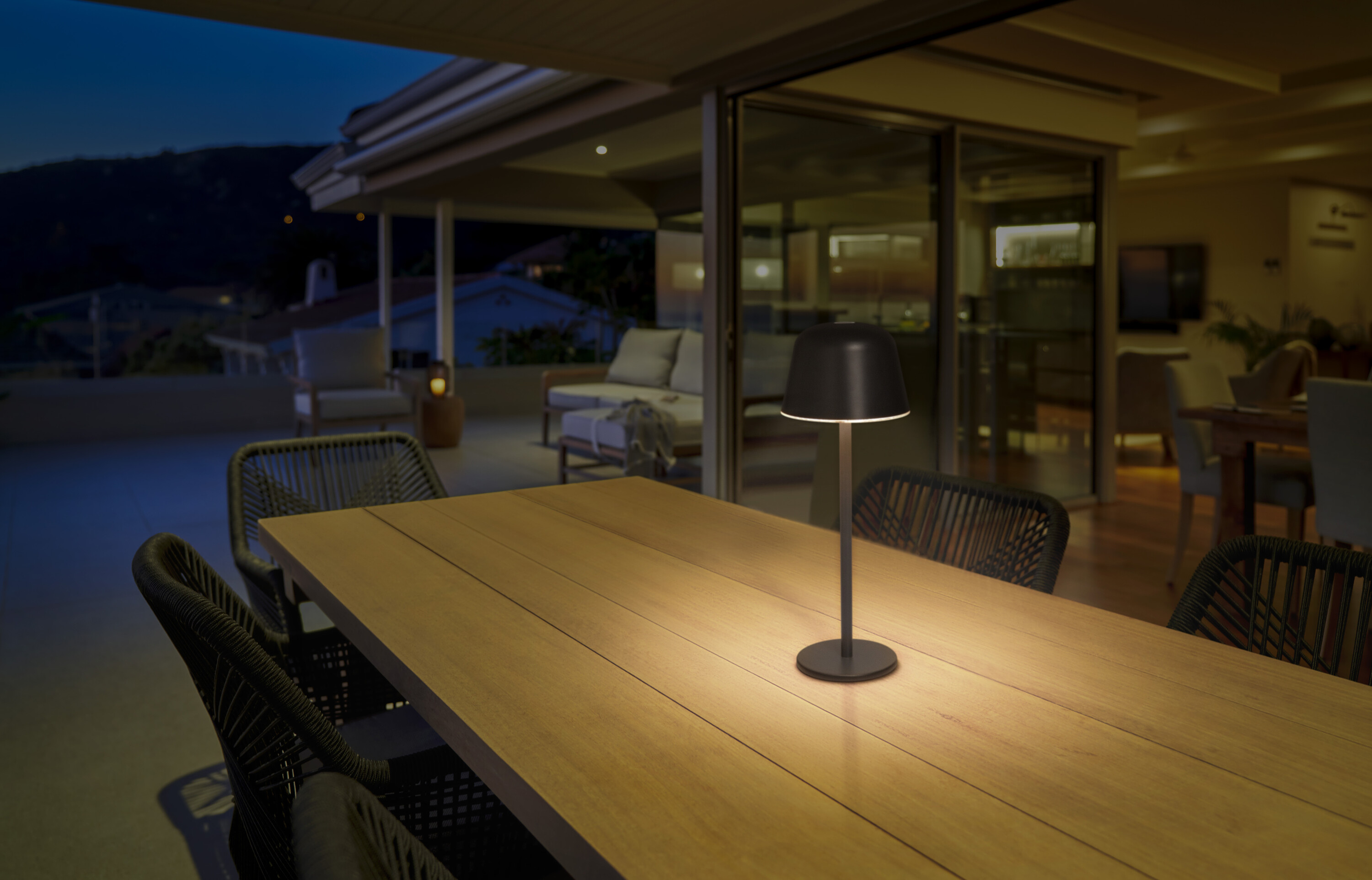 Scena all'aperto di notte con lampada da tavolo illuminata su un tavolo di legno, mobili da patio e cielo notturno.