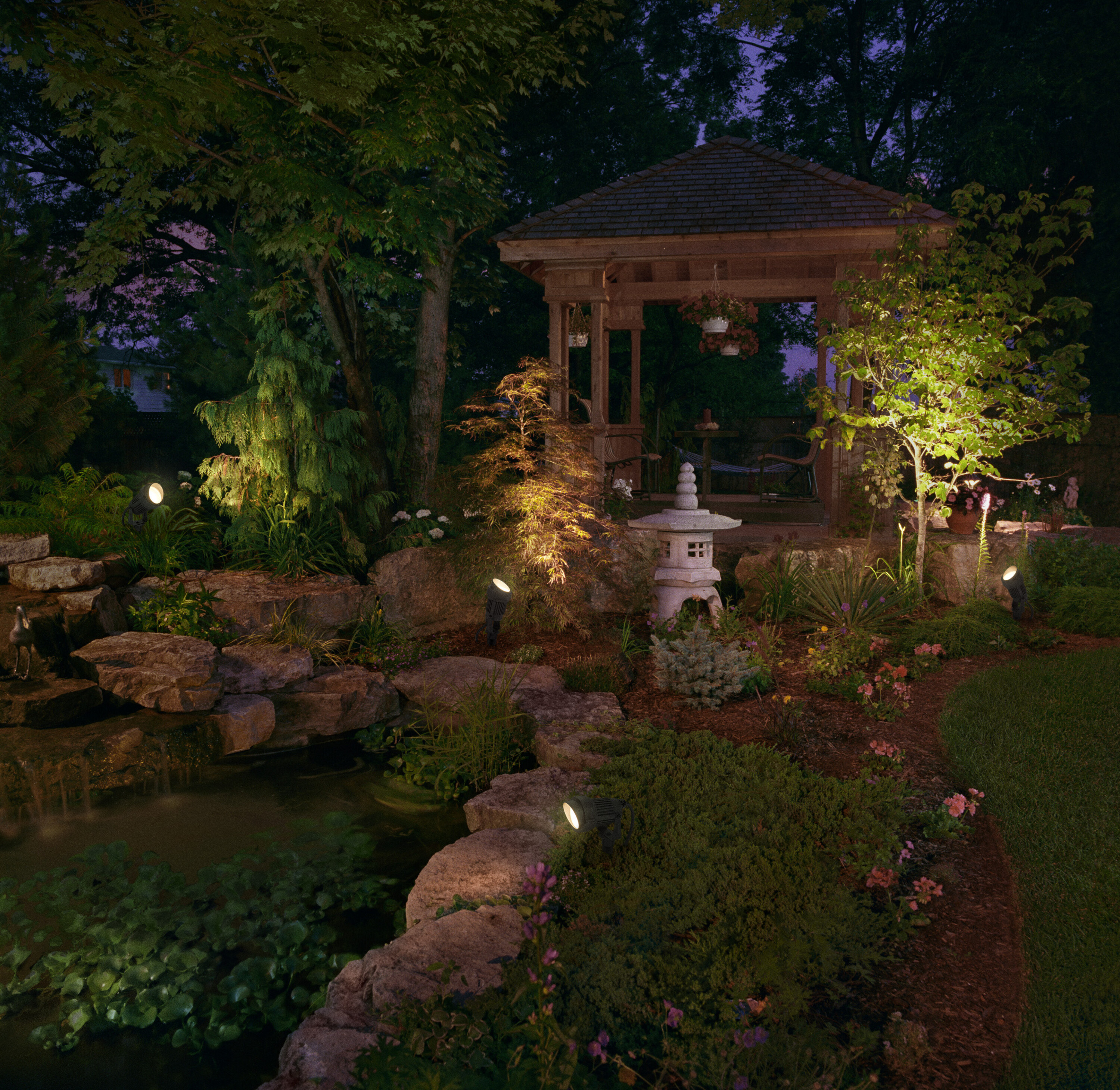 Eine Gartenszene bei Nacht mit Lichtern, die einen Pavillon, einen Teich und Bäume beleuchten. Felsen und Pflanzen sind sichtbar.