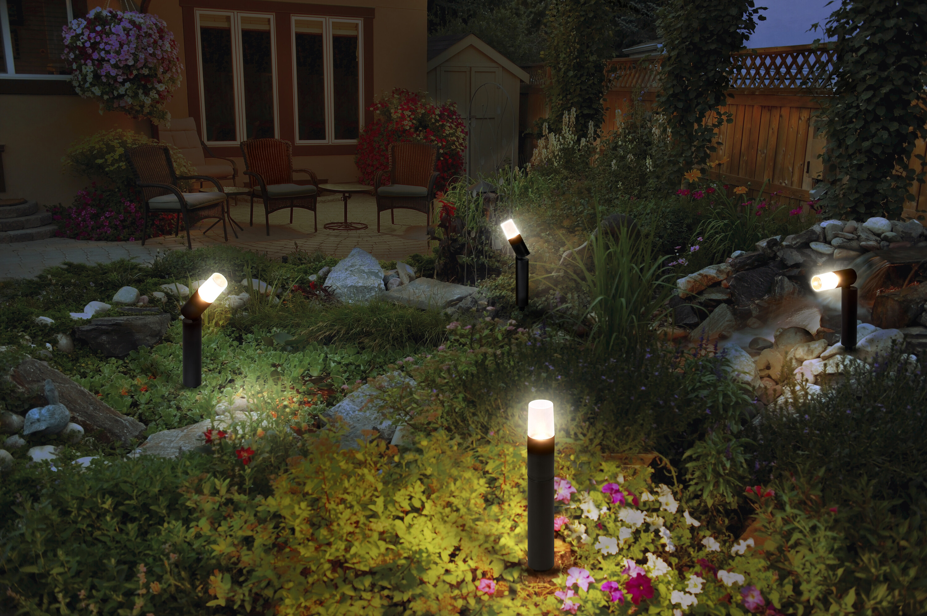 Garten in der Dämmerung, beleuchtet von Lampen. Pflanzen und ein Steinweg umgeben die Lampen, mit einem Haus im Hintergrund.