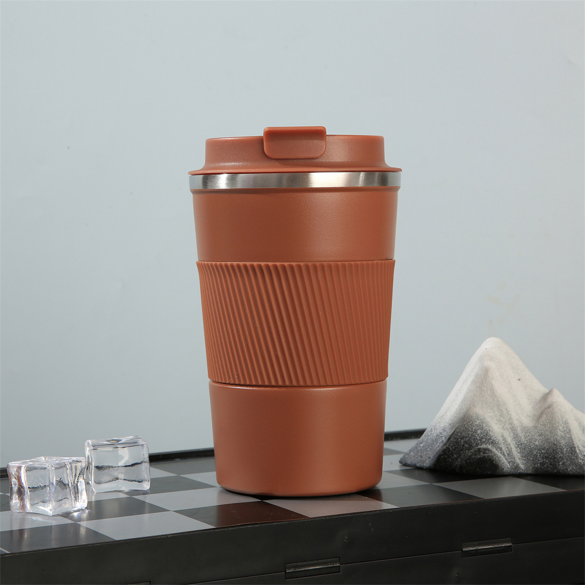Ein brauner Reisebecher mit geripptem Band und silbernem Deckel, neben Eiswürfeln und einem Teebeutel.