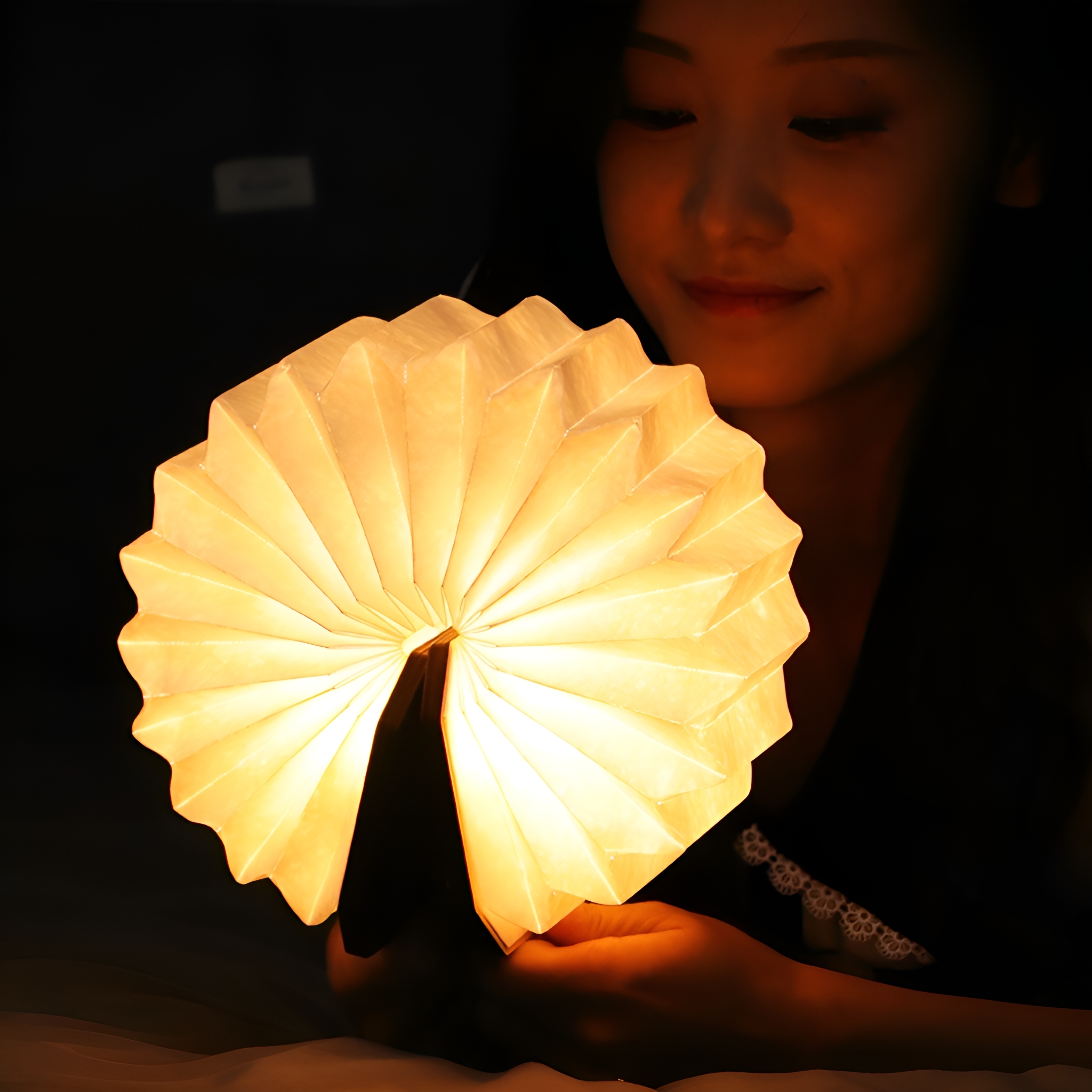 Eine Frau hält eine leuchtende, weiße Papierlampe. Die Lampe hat die Form eines Fächers. Gedämpfte Beleuchtung.