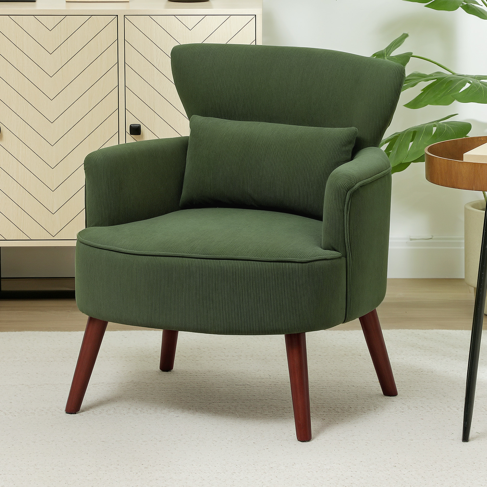 Poltrona verde con cuscino. Credenza in legno e pianta sullo sfondo. Tappeto bianco sotto la sedia.