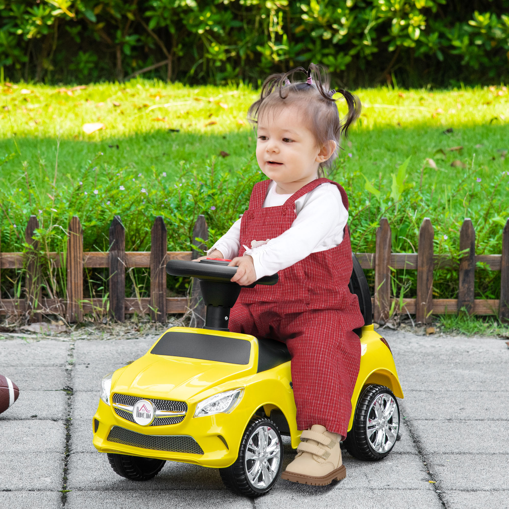 Un bambino è seduto in un'auto giocattolo gialla. Indossa una tuta rossa e una maglia bianca. Erba verde e una recinzione sullo sfondo.