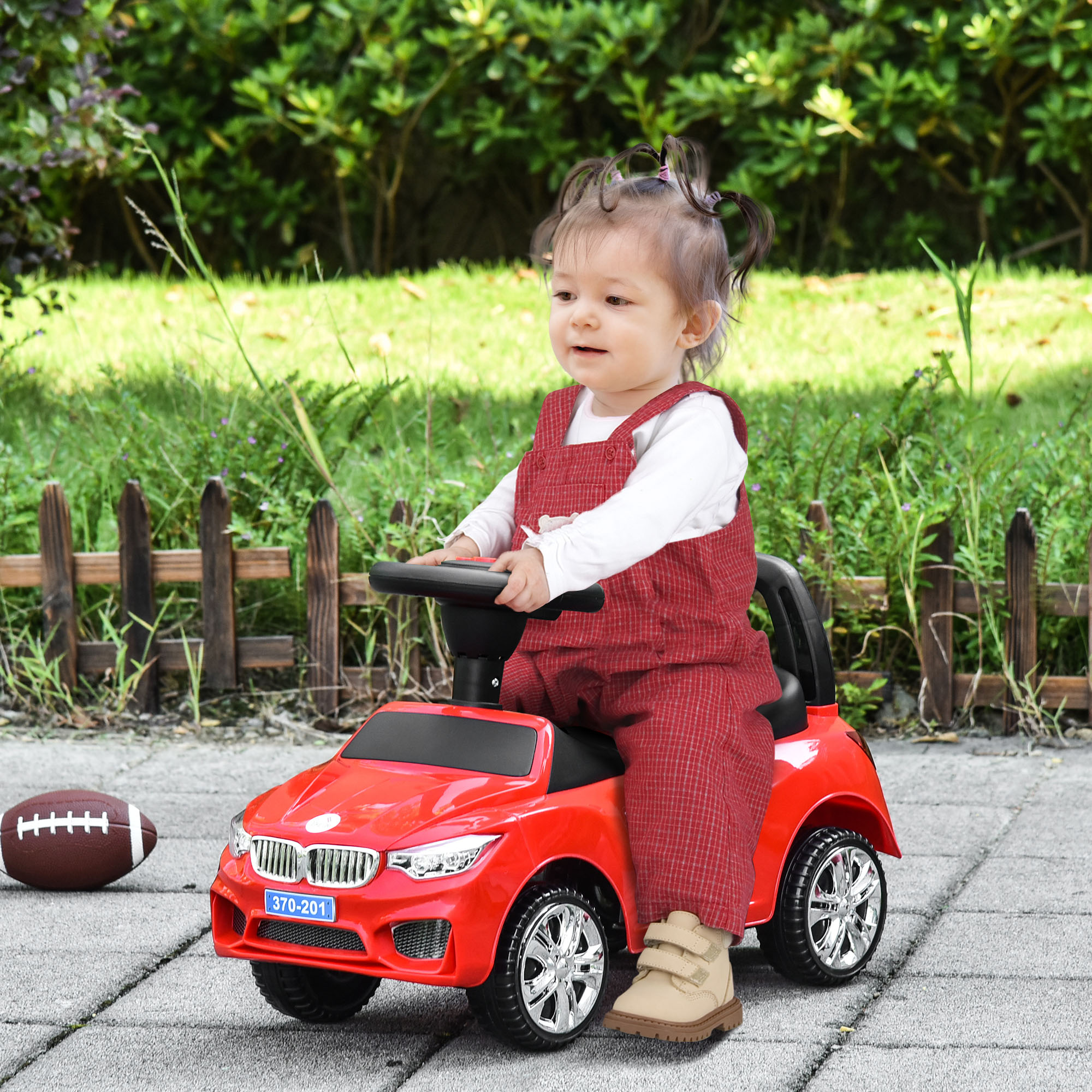 Un bambino con tuta rossa guida un'auto giocattolo rossa. Un pallone da calcio è a terra.