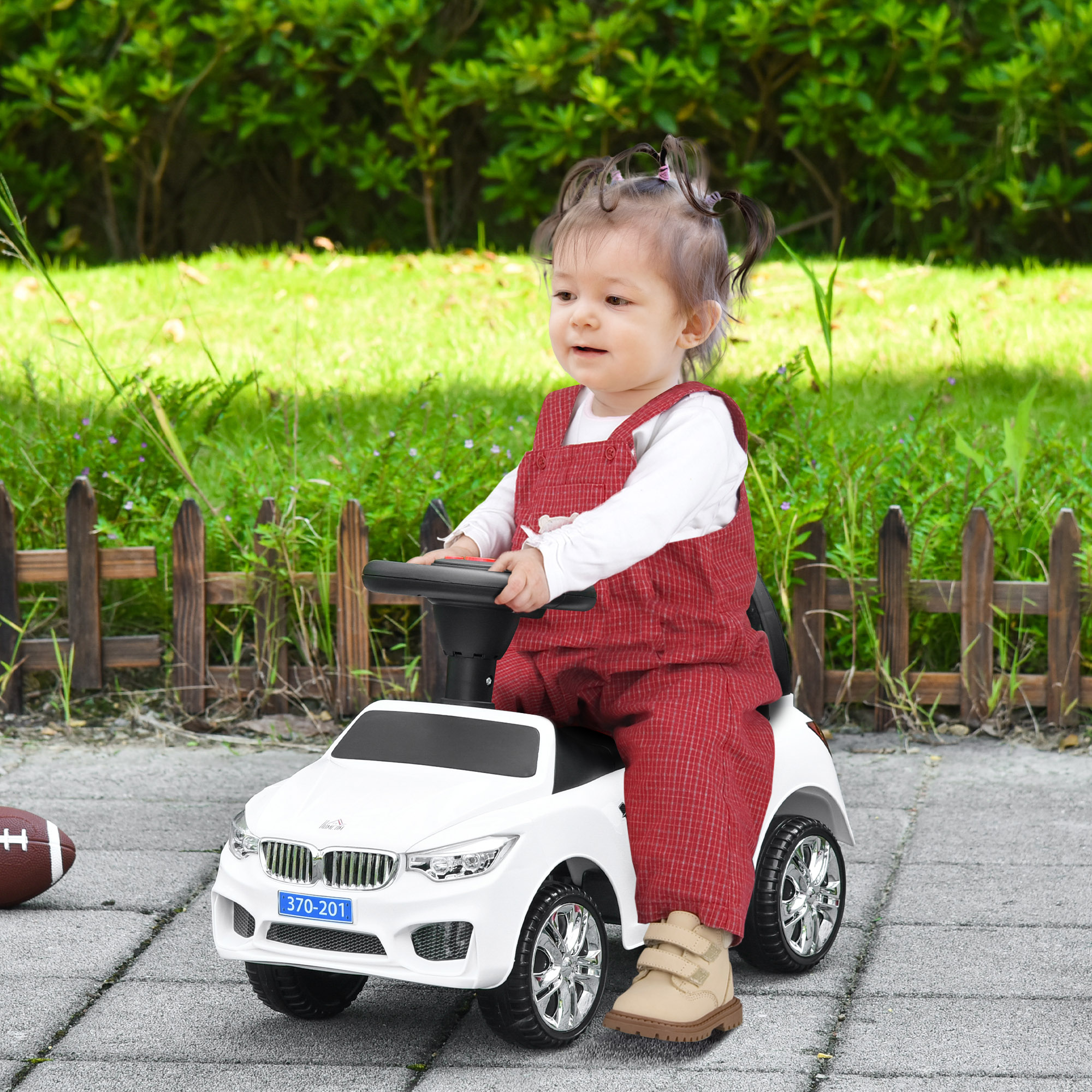 Un bambino è seduto in un'auto giocattolo bianca. Indossa una tuta rossa e una maglia bianca. Erba verde e una recinzione sullo sfondo.