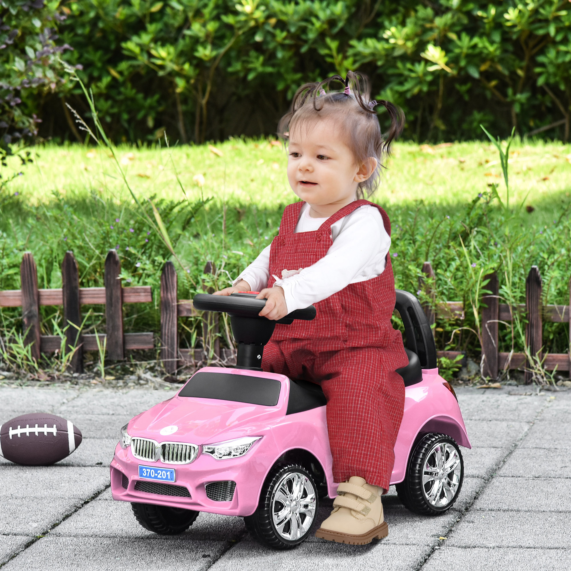 Una ragazzina con una tuta rossa guida un'auto giocattolo rosa su un sentiero di pietra. C'è un pallone da football.