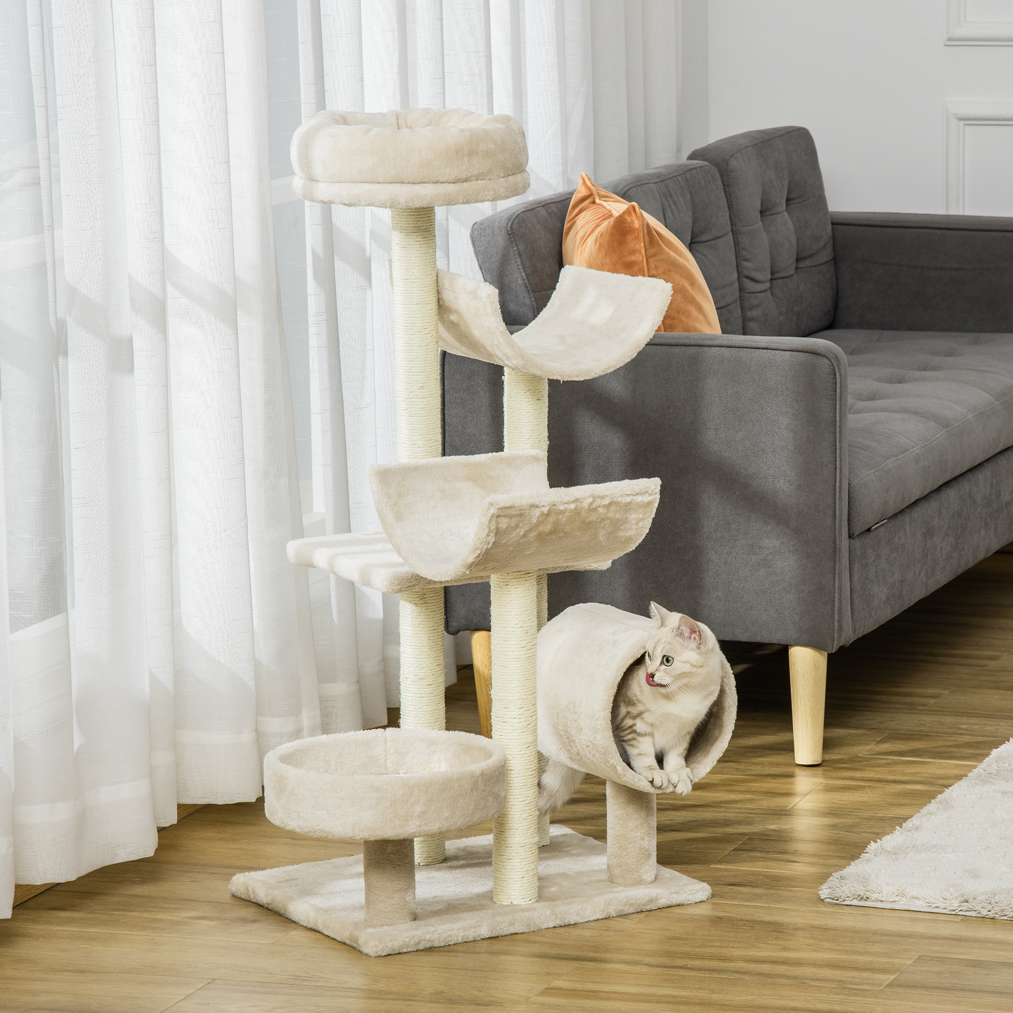 Árbol para gatos con múltiples niveles, una cama y un gato dentro de un tubo. Sofá gris y cortinas blancas en el BG.
