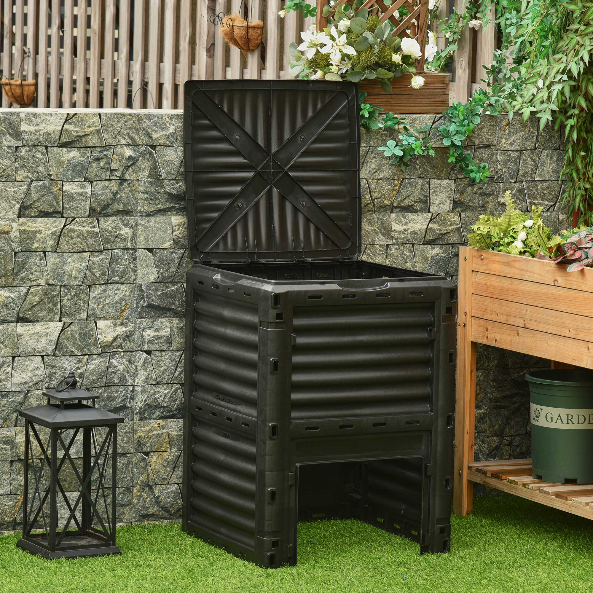 Bidone per compost nero con coperchio aperto, posto sull'erba con un muro di pietra e piante sullo sfondo.
