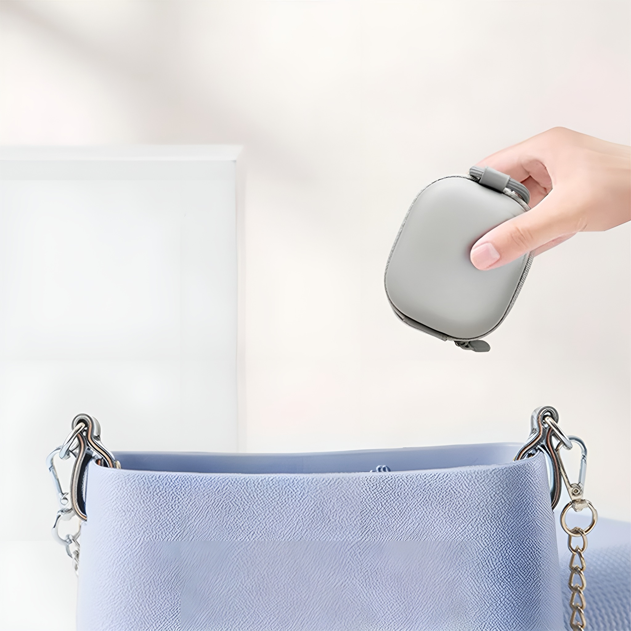 Eine Hand, die einen grauen Koffer in eine blaue Handtasche legt. Die Handtasche hat silberne Haken.