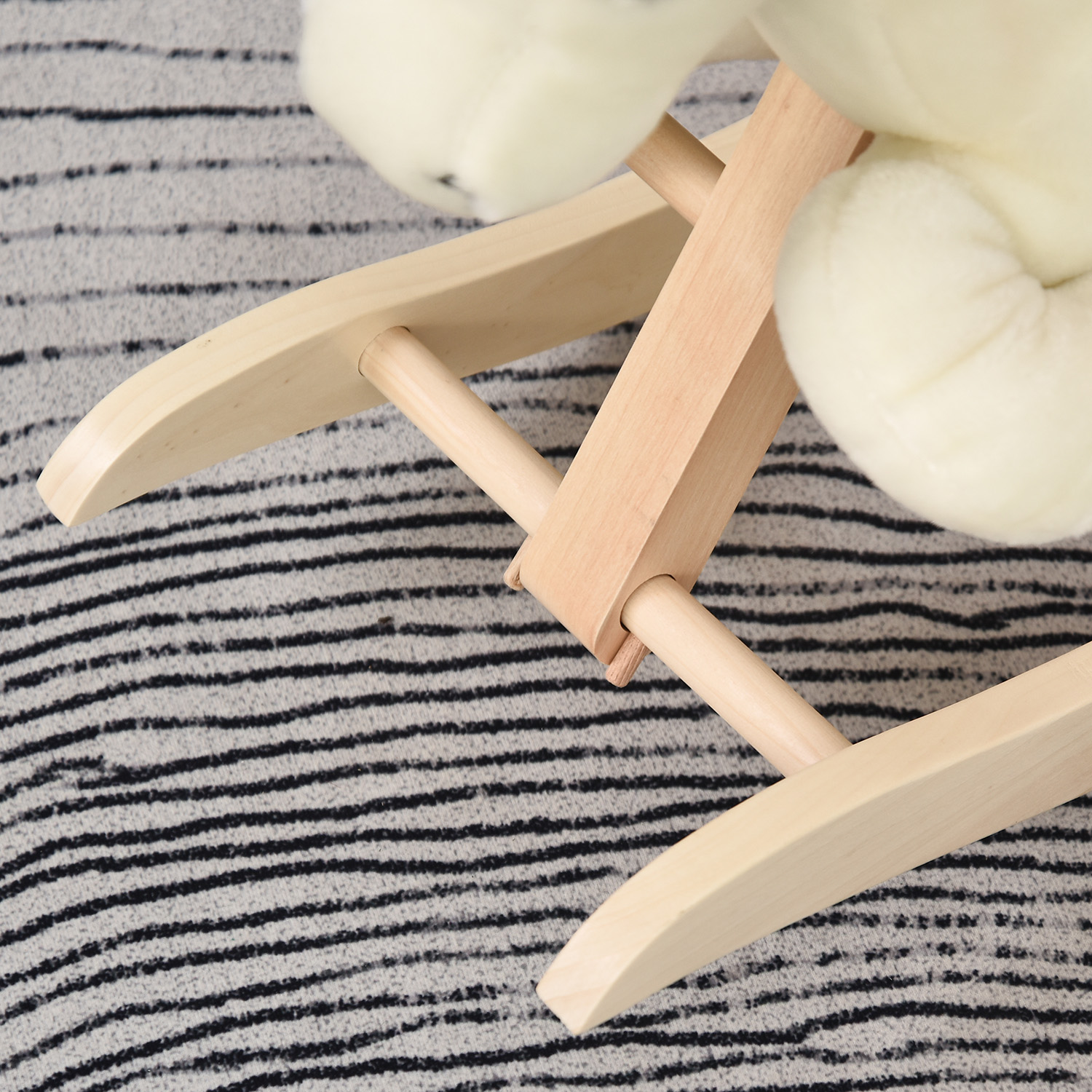 Base di un giocattolo a dondolo in legno chiaro con un peluche bianco su un tappeto grigio a righe.