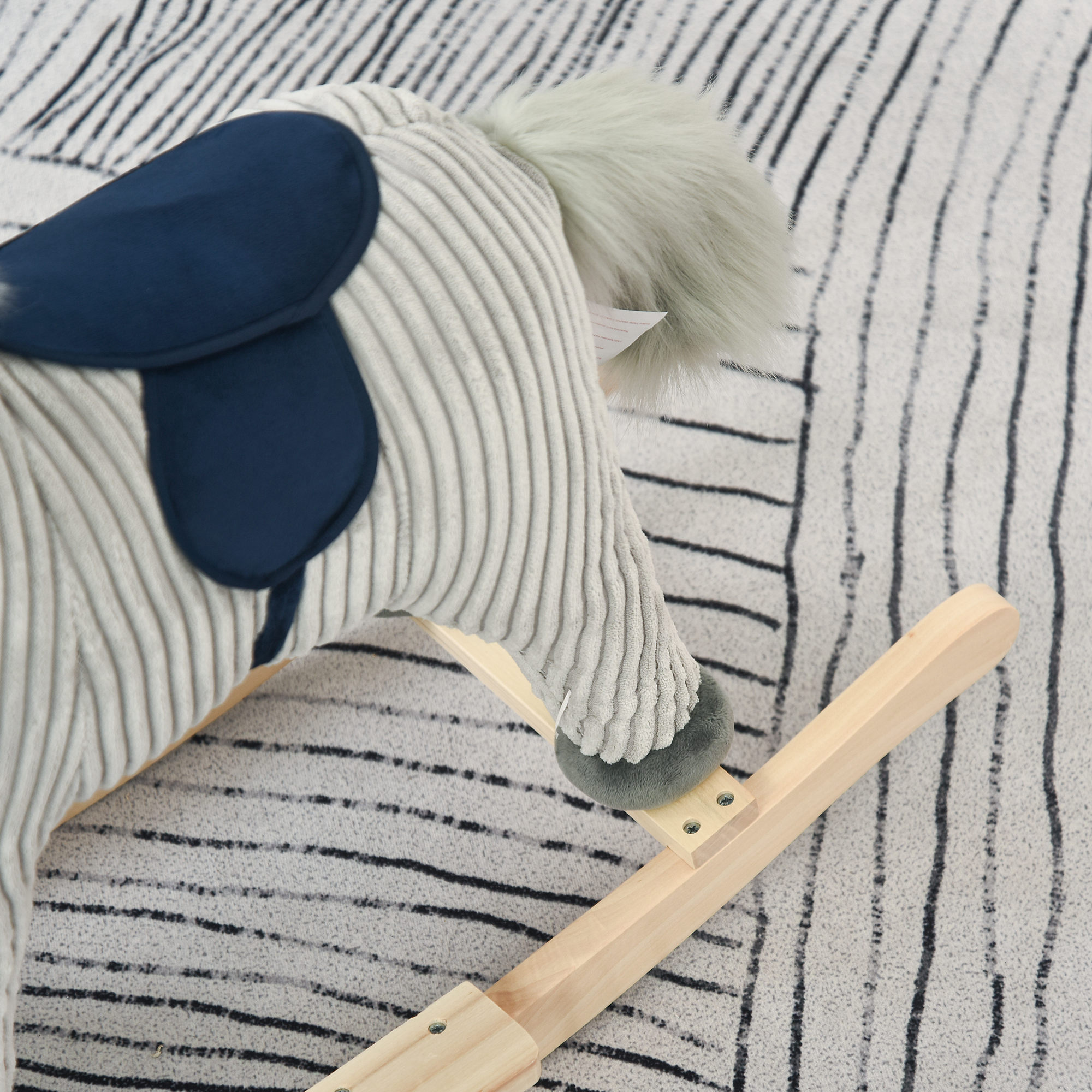 Primo piano della testa di un cavallo a dondolo grigio e bianco. Sella blu. Il cavallo è su un bilanciere di legno. Lo sfondo è un tappeto.