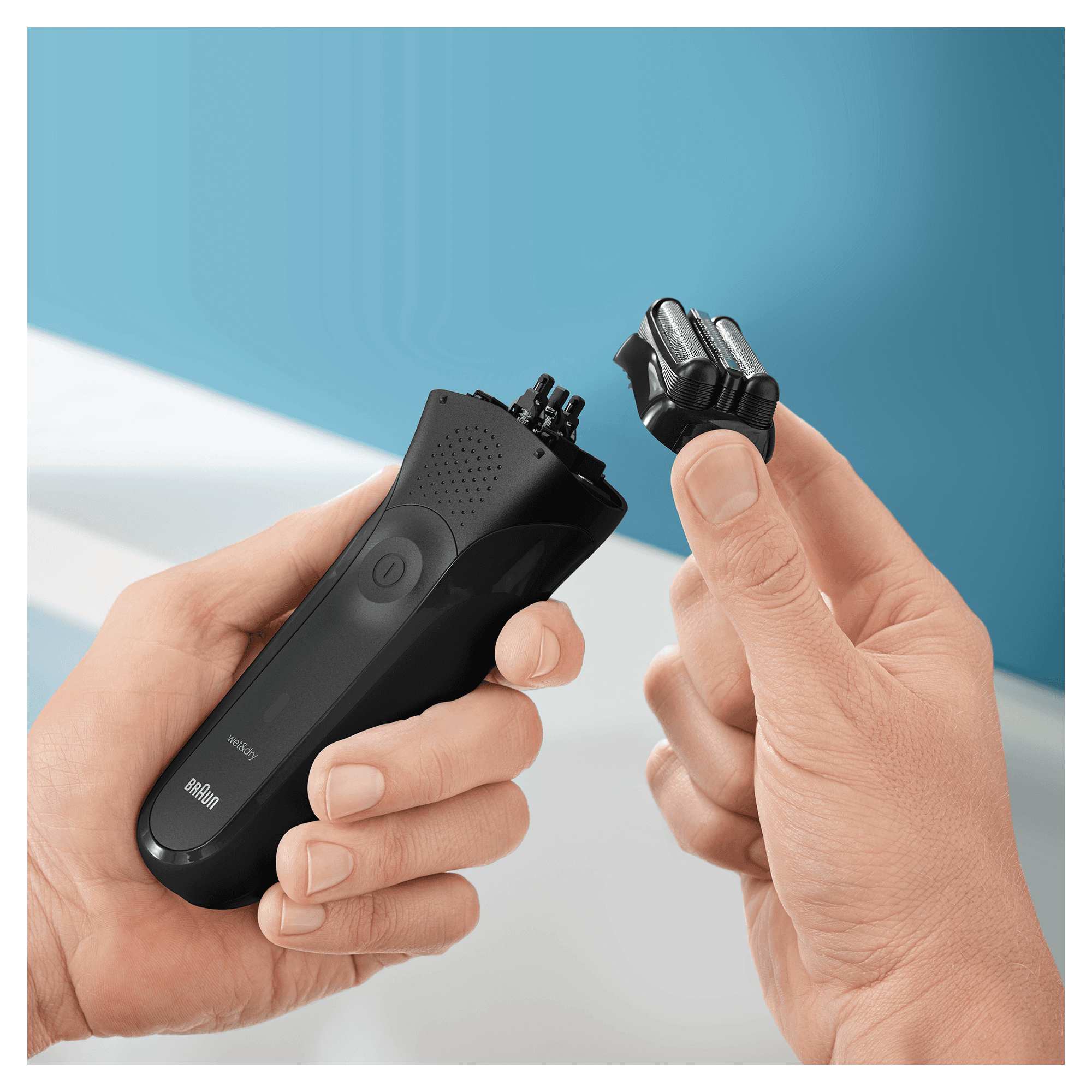 Dłonie trzymające czarną golarkę elektryczną, z odłączoną głowicą, na niebieskim tle.