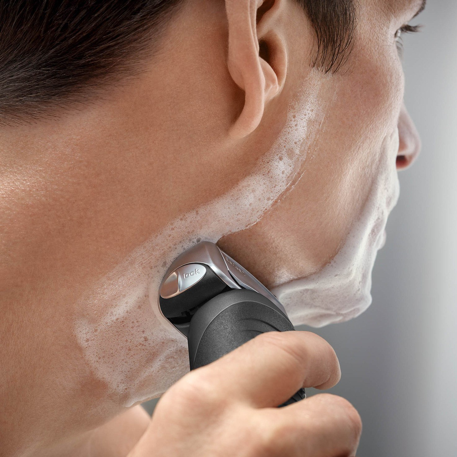 Mężczyzna goli twarz pianką za pomocą srebrnej i czarnej golarki elektrycznej. Golarka jest w użyciu.