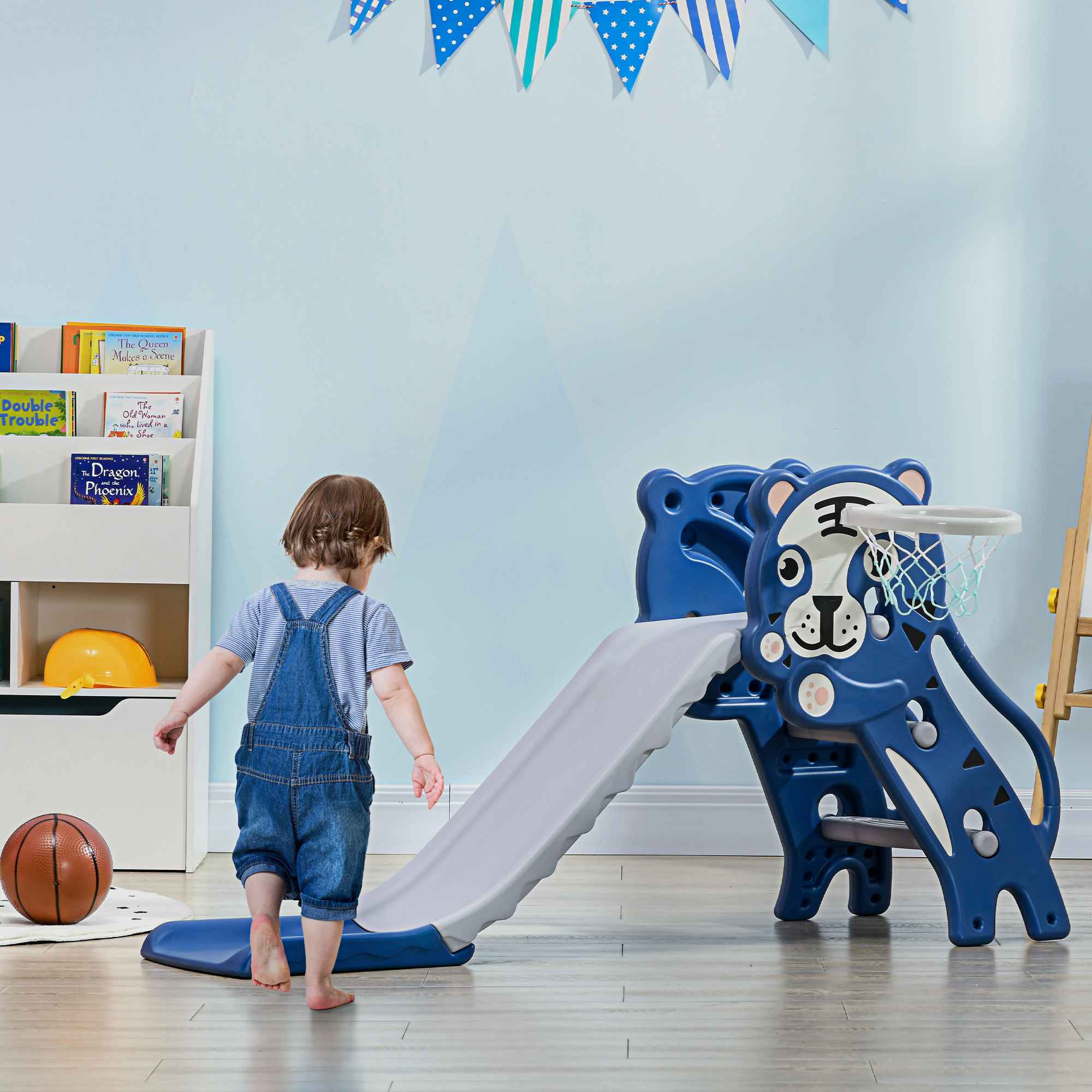 Un bambino cammina verso uno scivolo blu in una stanza con una libreria e uno striscione.