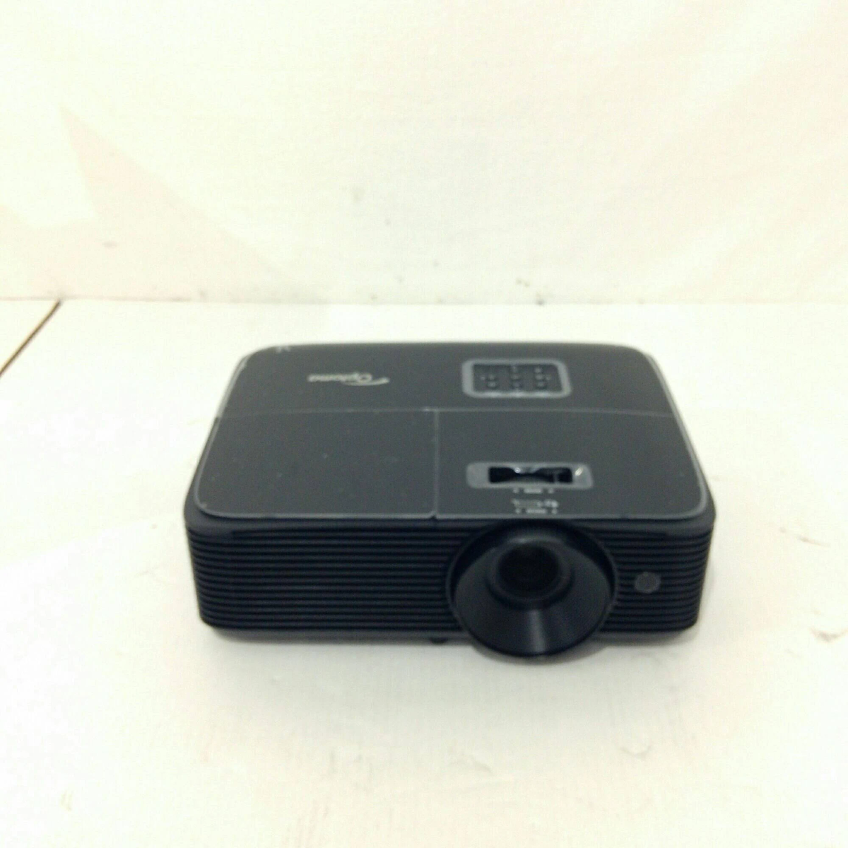 Czarny projektor Optoma na białej powierzchni, z obiektywem z przodu.