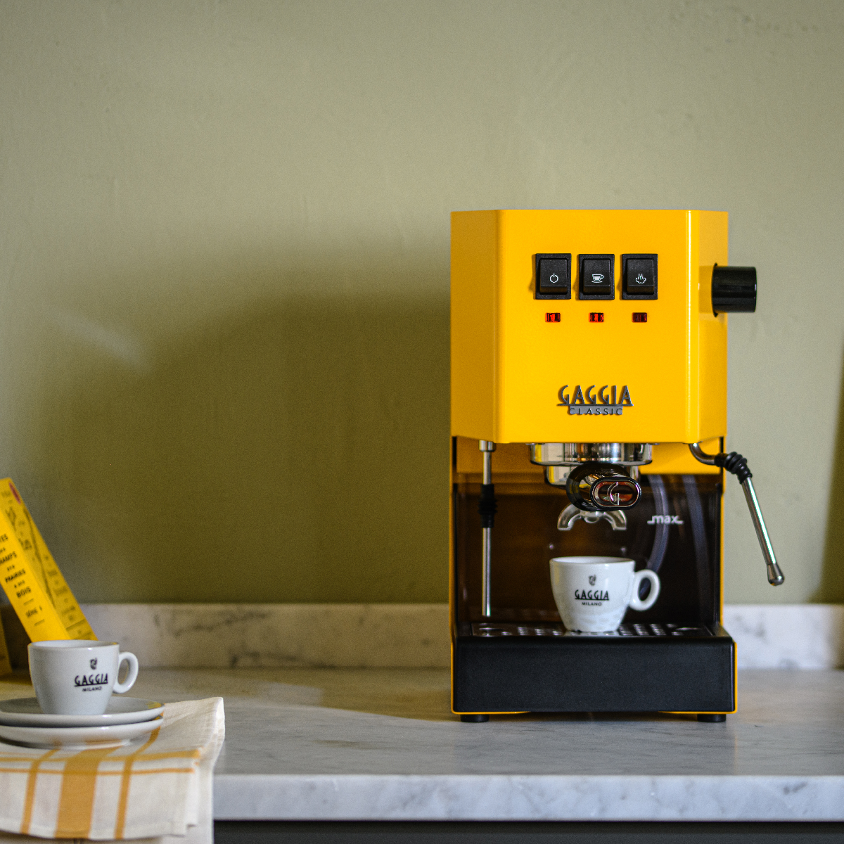 Beyaz tezgah üzerinde iki fincanlı sarı bir Gaggia Classic espresso makinesi, yeşil duvar arka planı.
