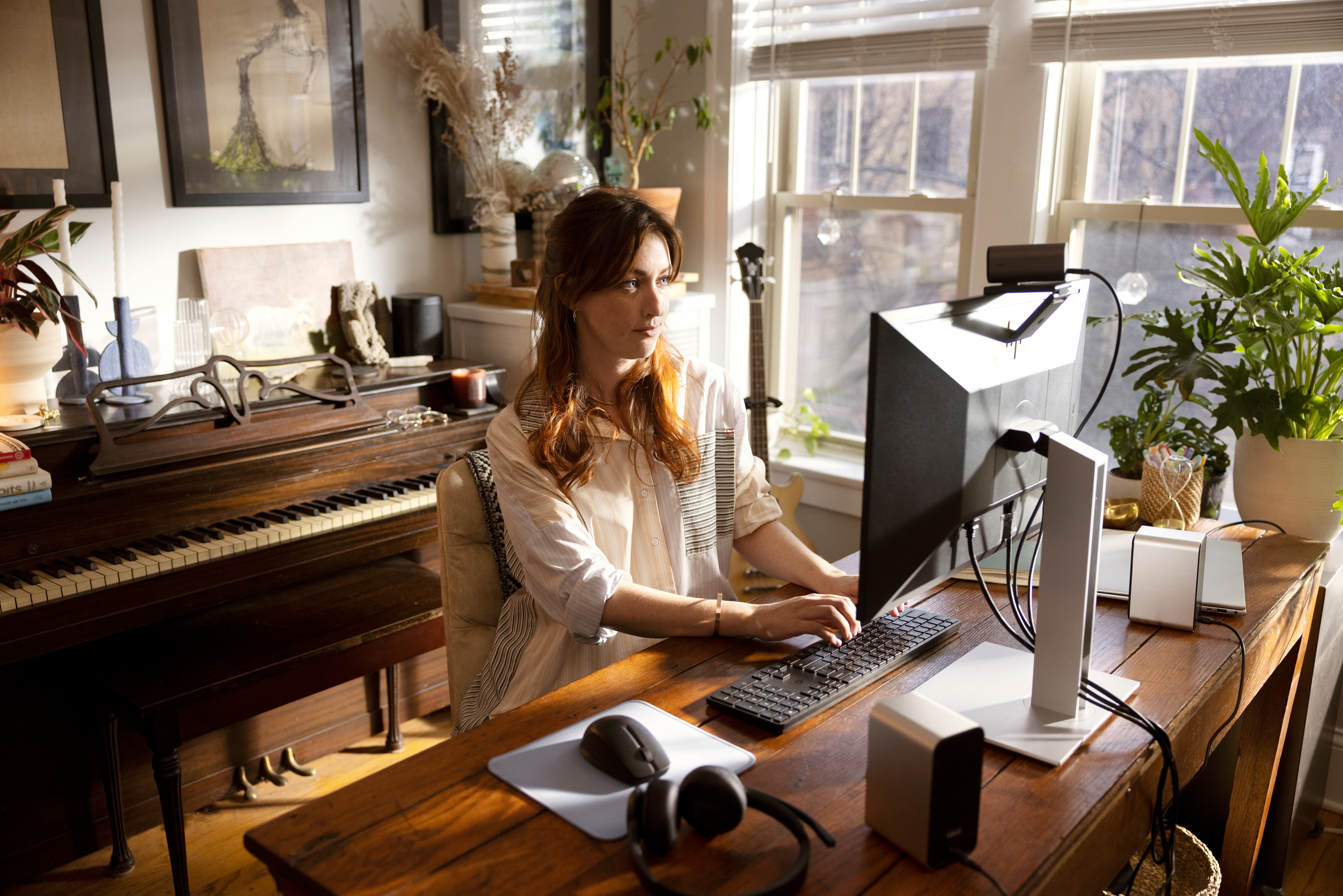 Kobieta przy biurku z komputerem, klawiaturą, myszą, słuchawkami, głośnikami, roślinami, pianinem w pokoju.