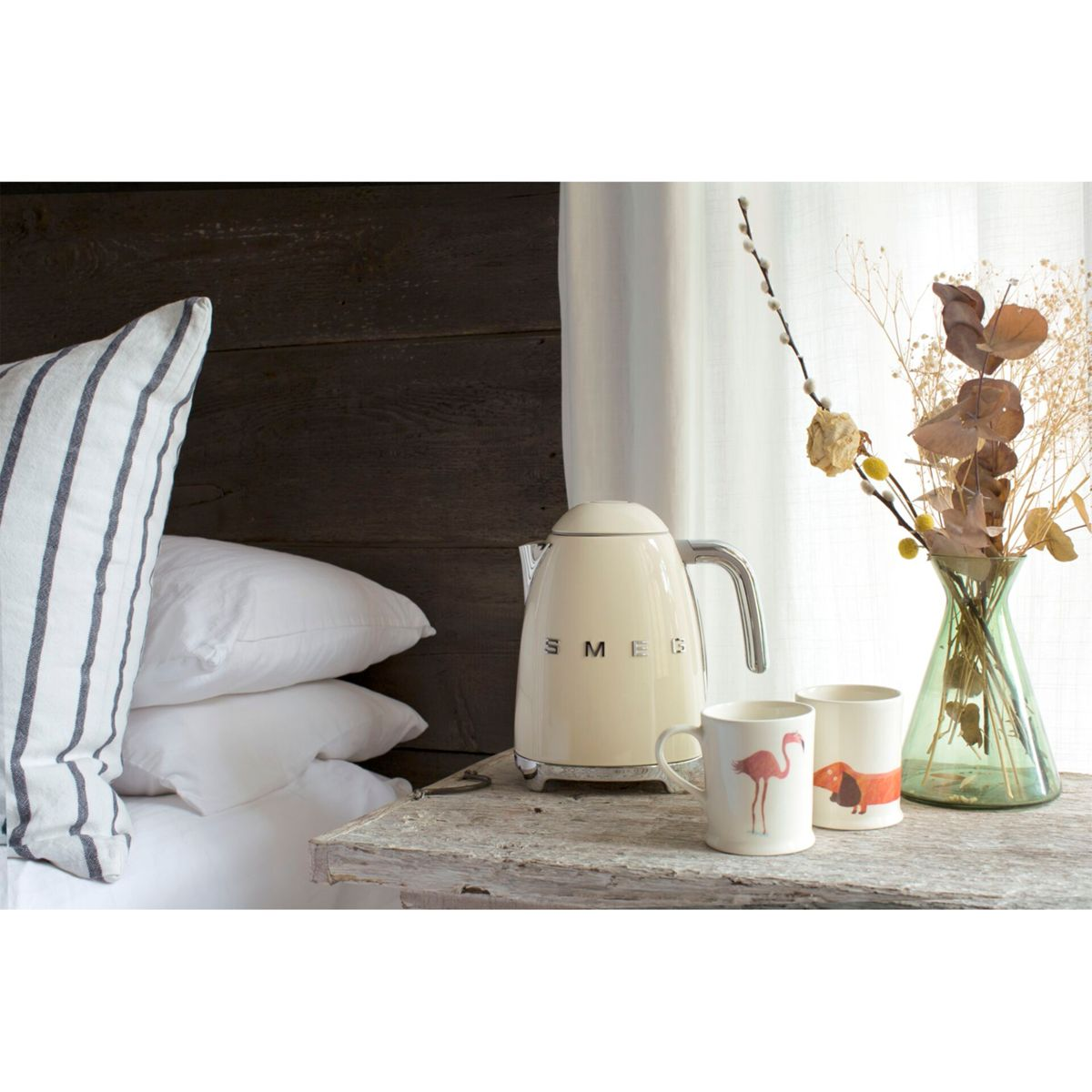 Beżowy czajnik Smeg i dwa kubki na drewnianym stole, z poduszkami i wazonem.