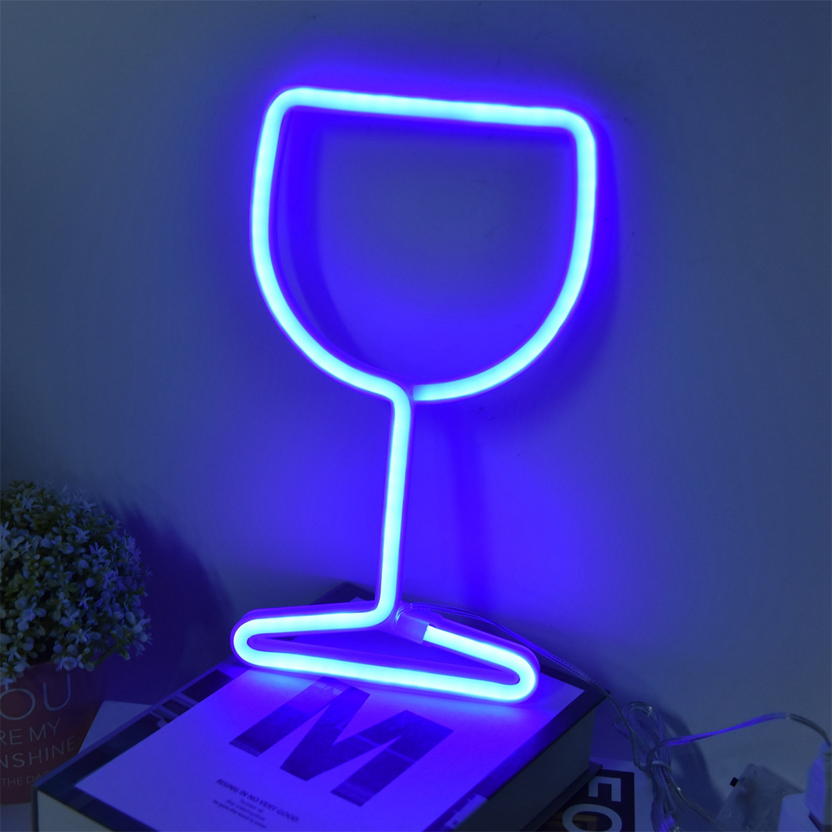 Ein blaues Neon-Weinglas auf einem Tisch mit einem Buch und kleinen Pflanzen. Der Hintergrund ist dunkelblau.