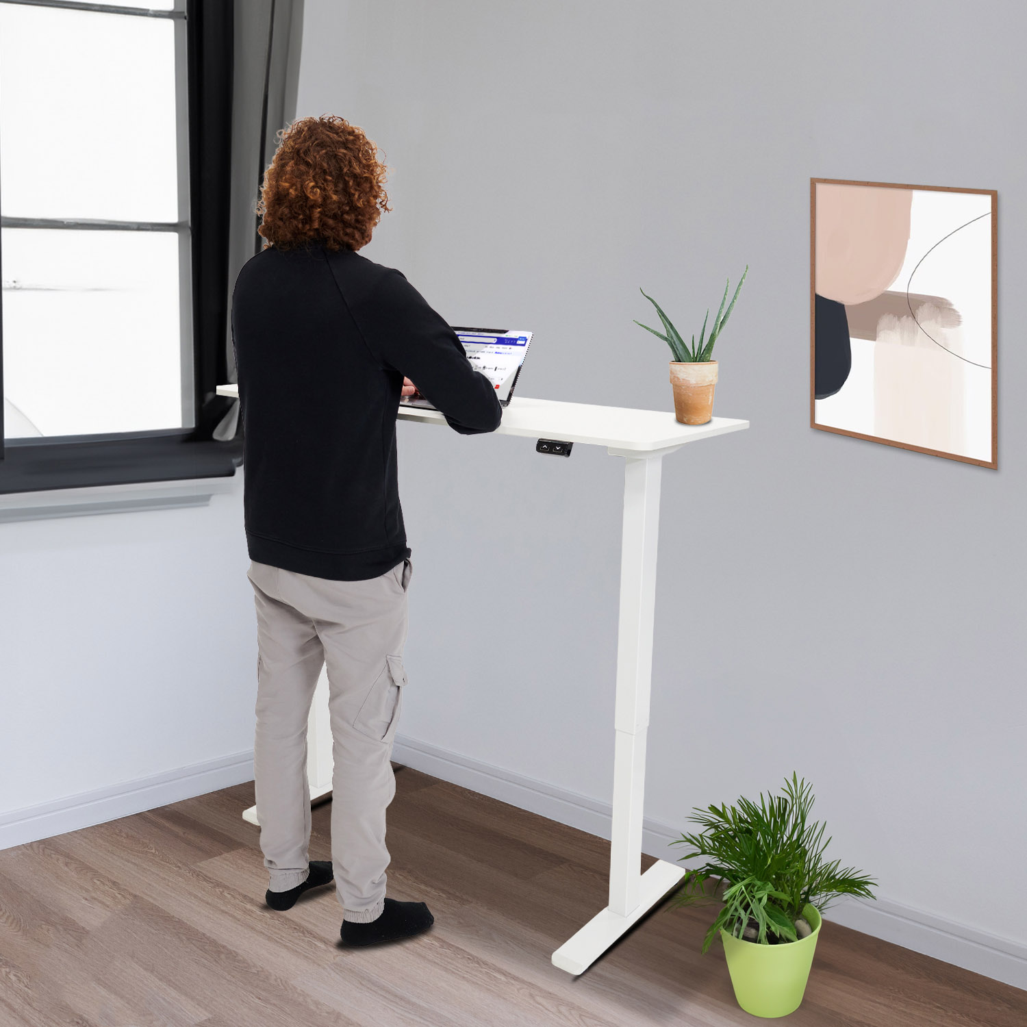 Een persoon staat bij een witte verstelbare bureau. Een laptop, plant en kunst zijn zichtbaar.