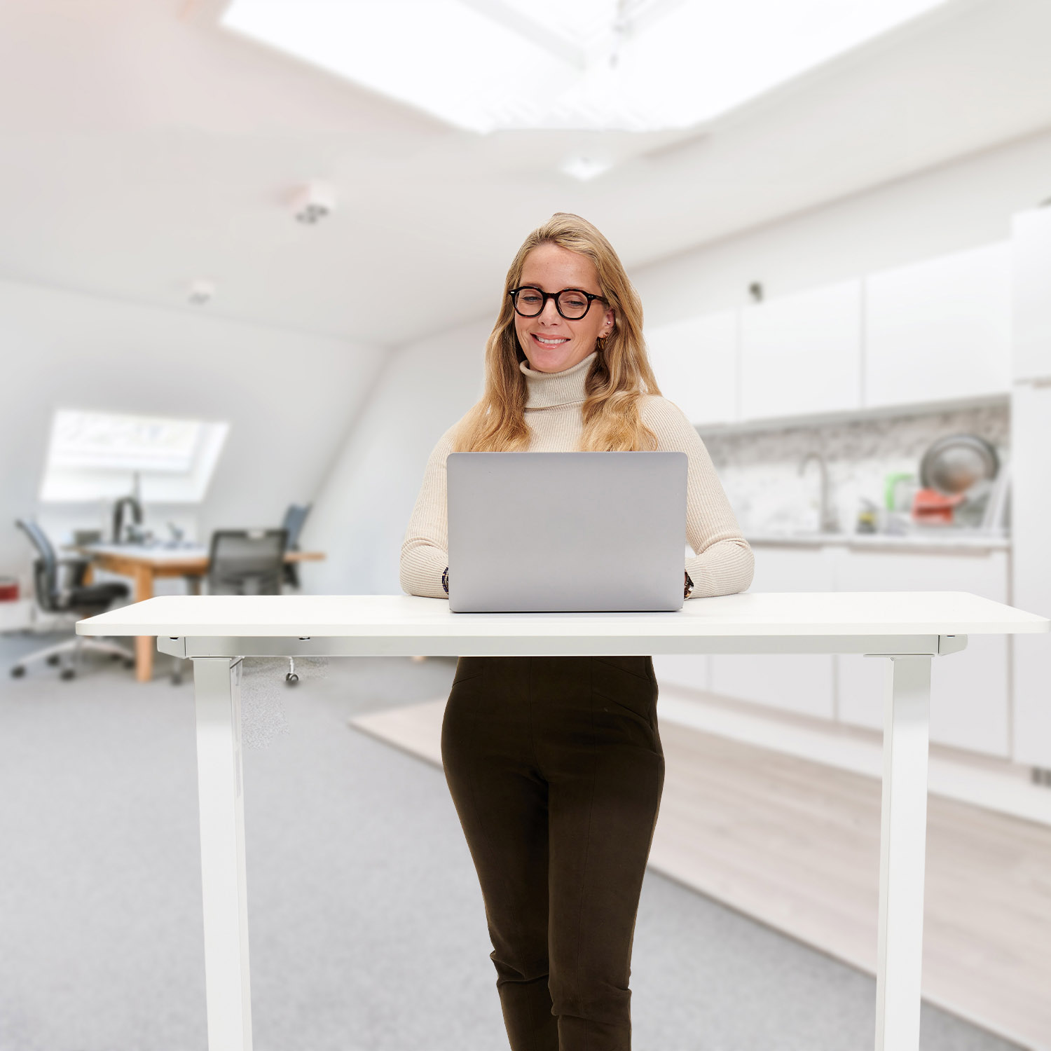 Vrouw gebruikt laptop aan stahoogte bureau. Witte trui, bril. Vage kantoorachtergrond.