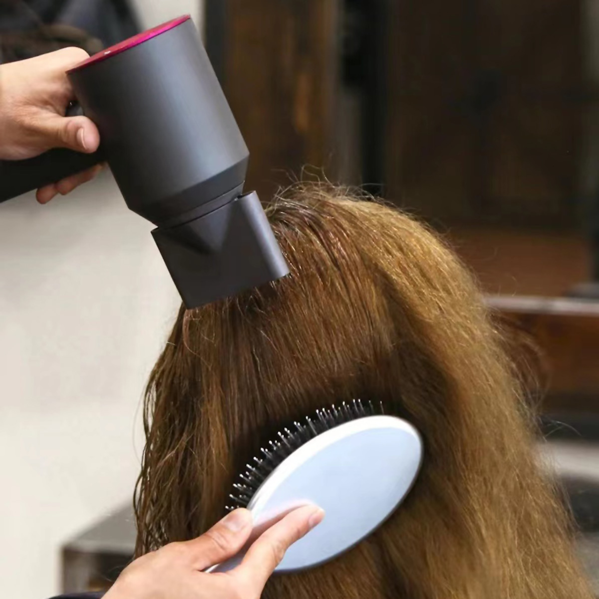 Mani usano asciugacapelli e spazzola su capelli castani. In un salone, con sfondo sfocato.