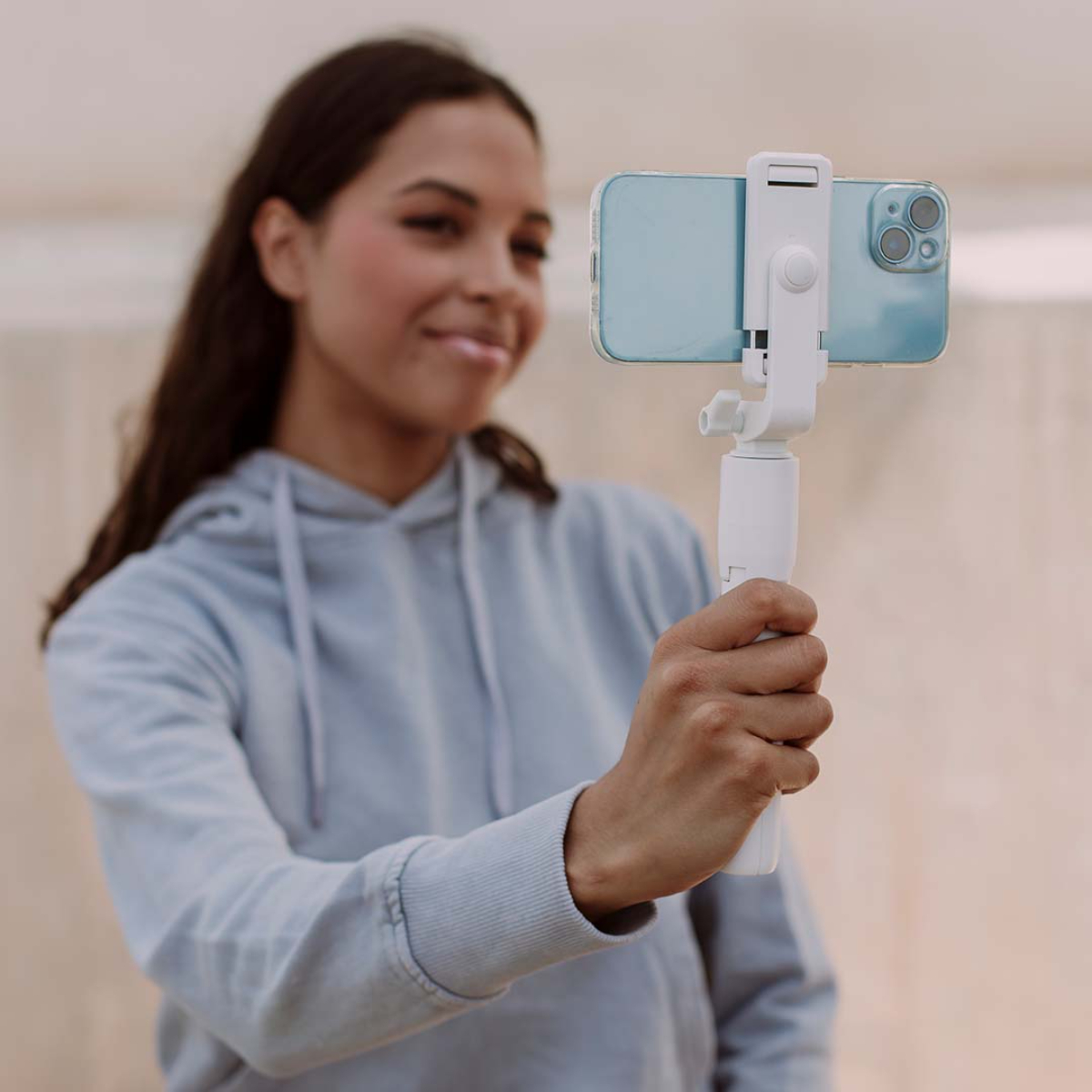 Una mujer sosteniendo un teléfono en un palo selfie, sonriendo, con un fondo claro.
