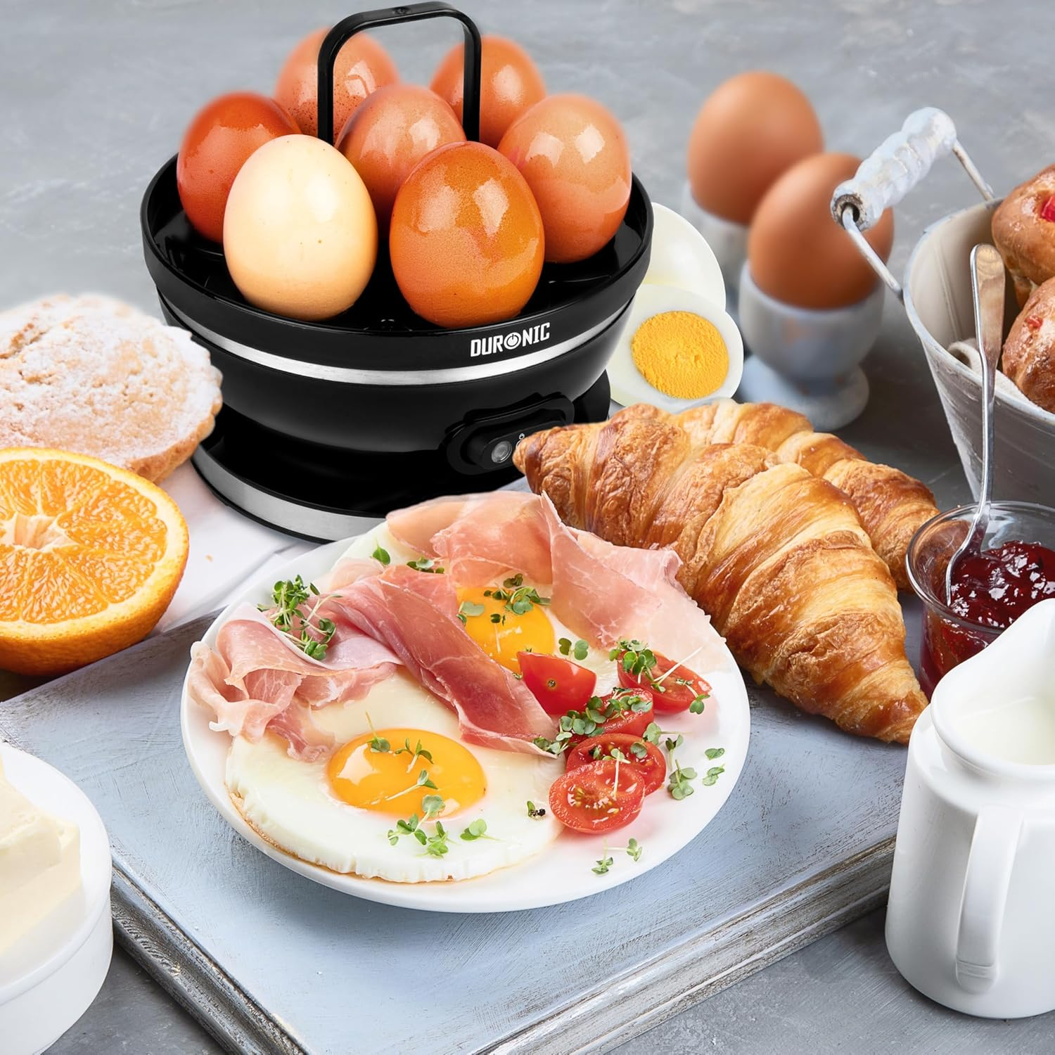 Ein Tisch mit Eierkocher, Eiern, Schinken, Croissants und anderen Frühstücksartikeln.