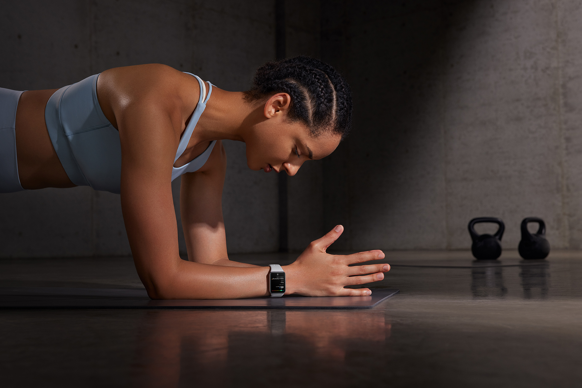 Kobieta w pozycji deski, nosząca smartwatch. Ciemne tło, dwie odważniki kulowe.