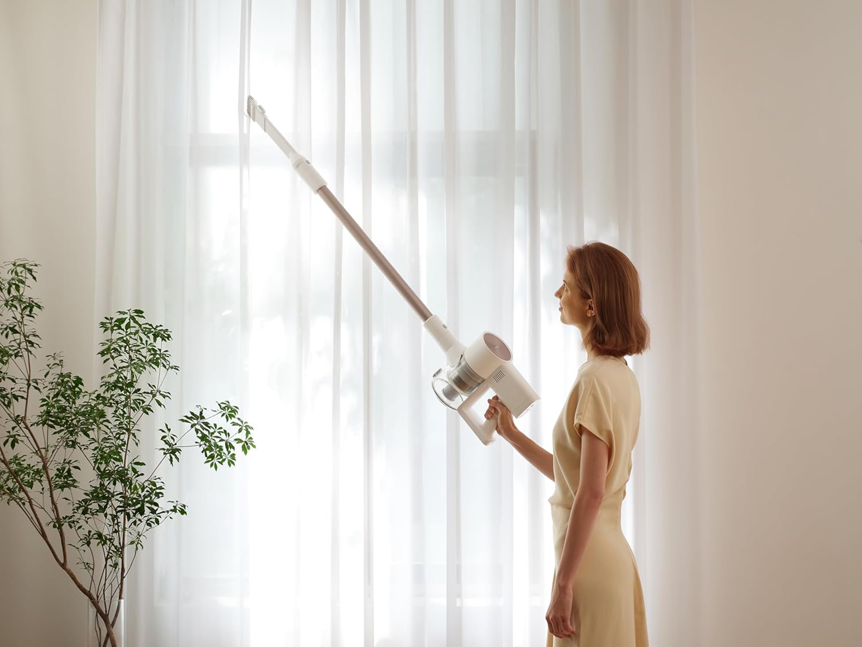 Kobieta odkurza w pobliżu okna z zasłonami. Trzyma biały, bezprzewodowy odkurzacz.