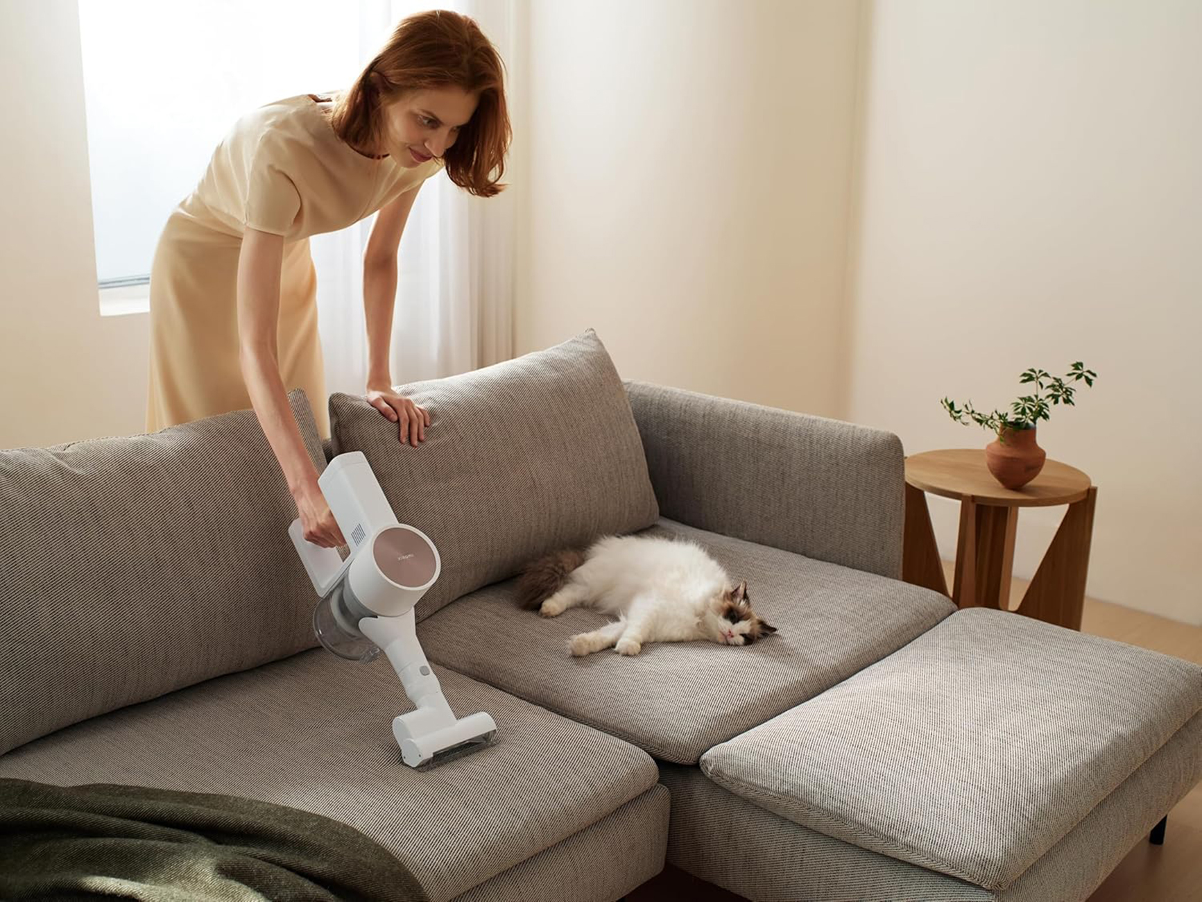 Kobieta odkurza sofę z kotem. Beżowa sofa i drewniany stolik.